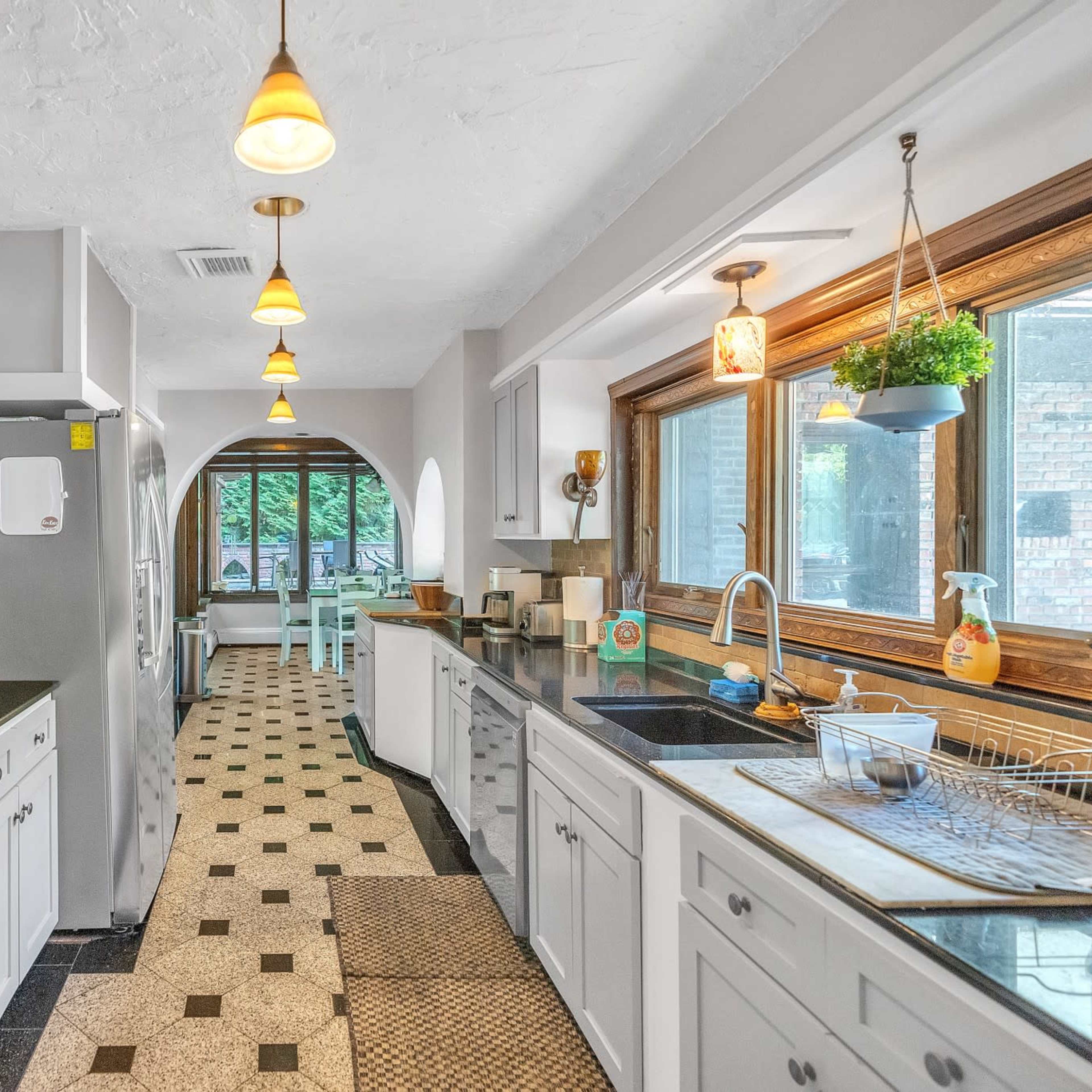 The kitchen features a long layout with dark countertops, white cabinetry, and multiple pendant lights, leading to a windowed area with light-colored furniture.