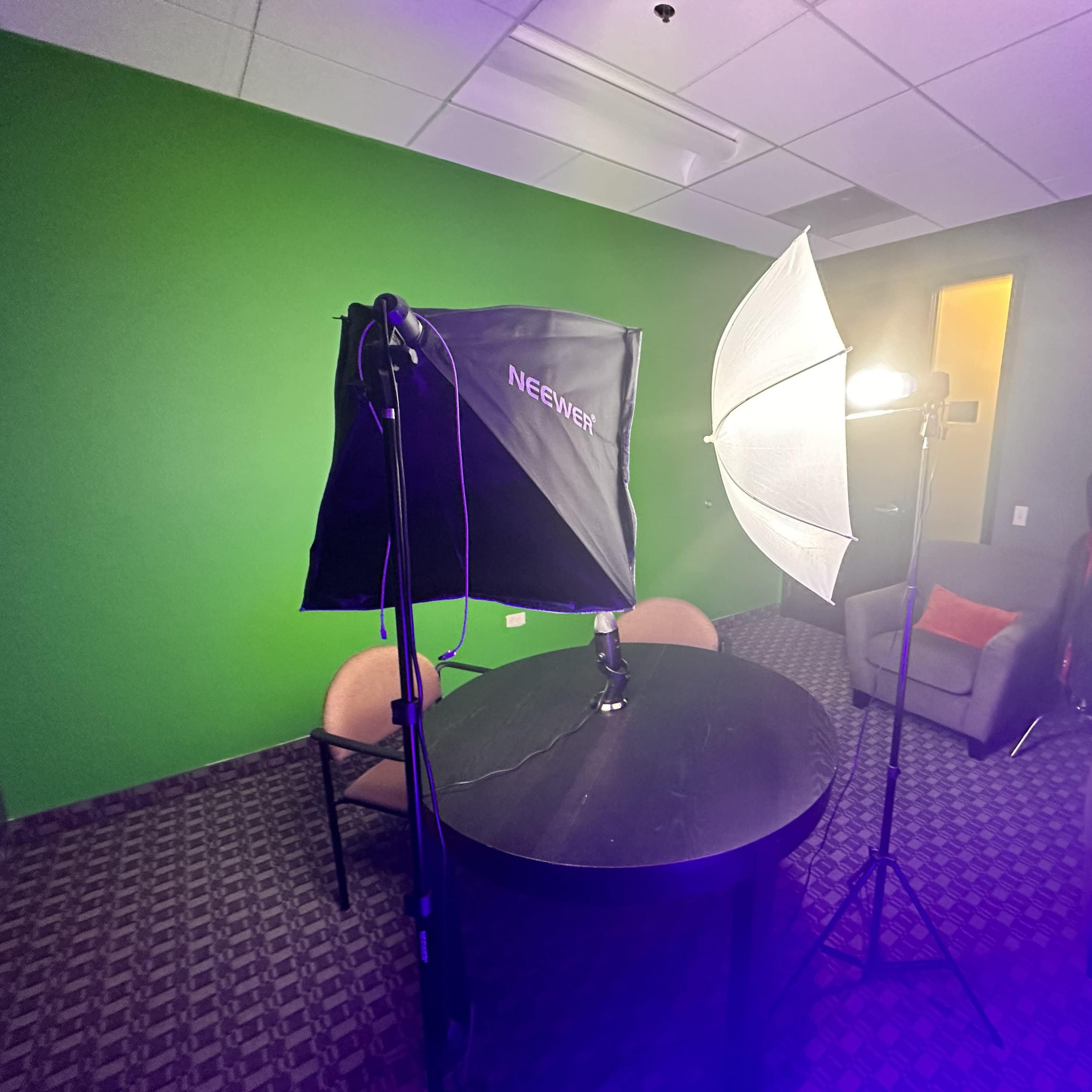 A small room with a round table, two chairs, and lighting equipment set up against a green screen backdrop.