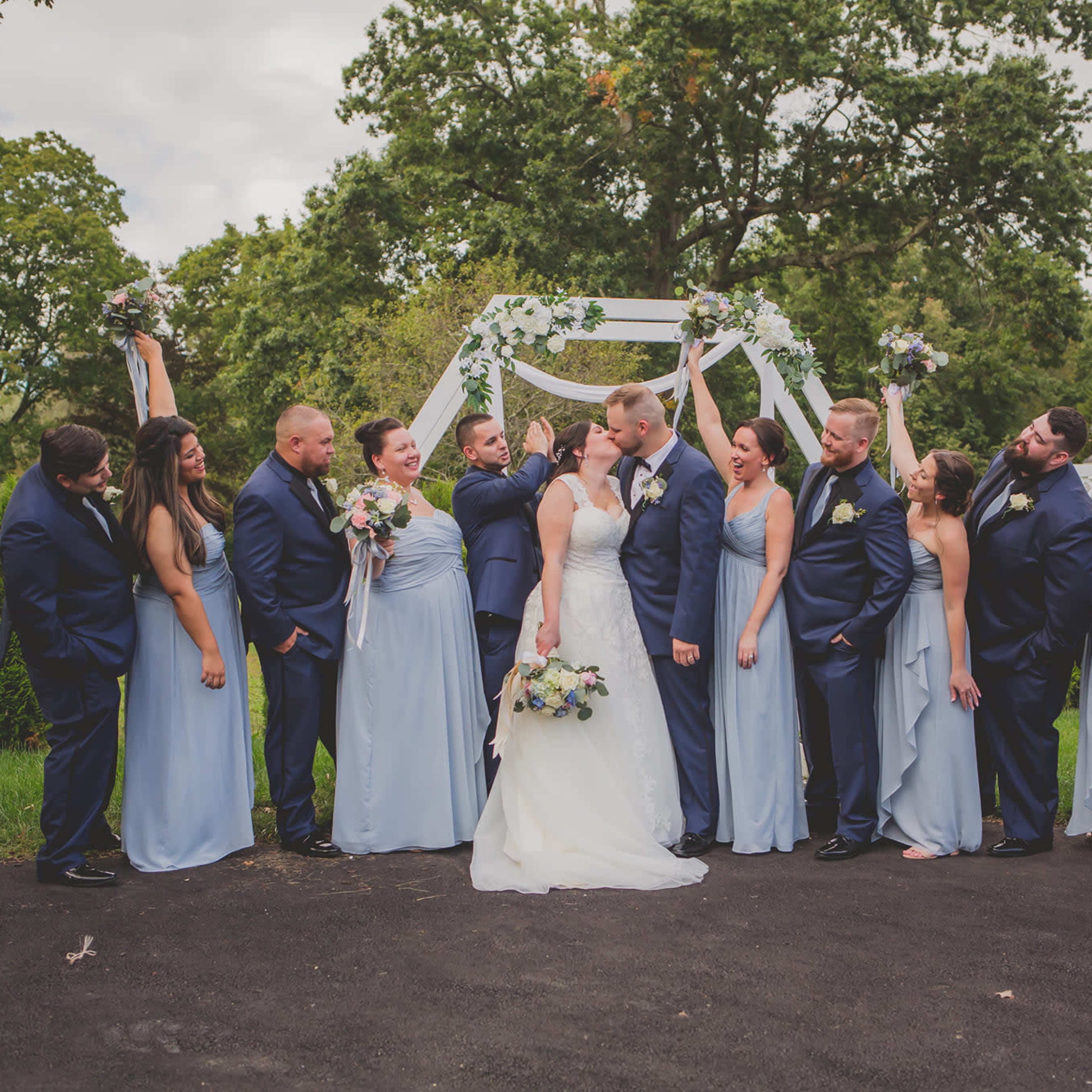 A bridal party in formal attire celebrates under a floral arch on a grassy venue.