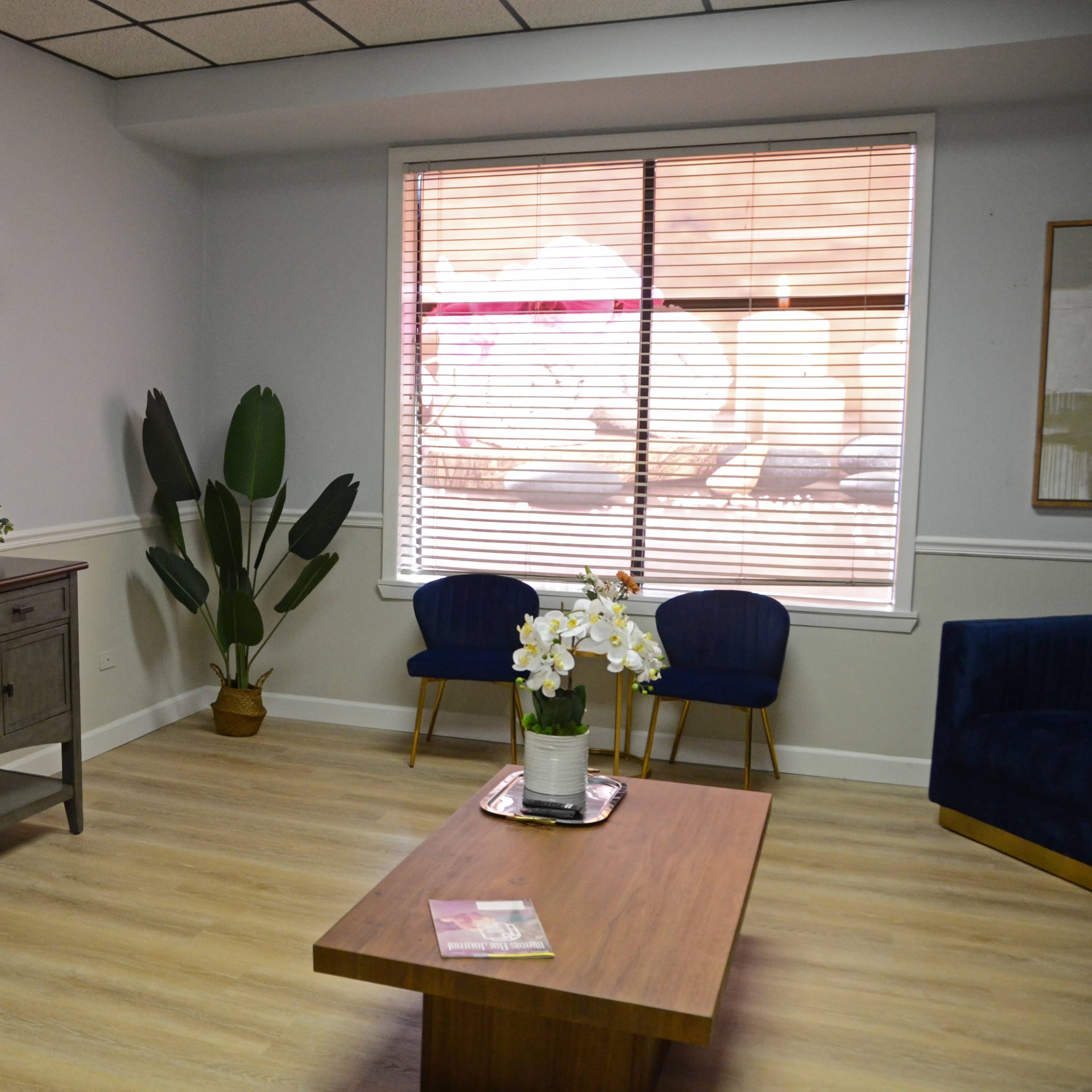 A modern waiting room with a wall-mounted TV, a wooden coffee table, a plant, and two blue upholstered chairs near a window with closed blinds.