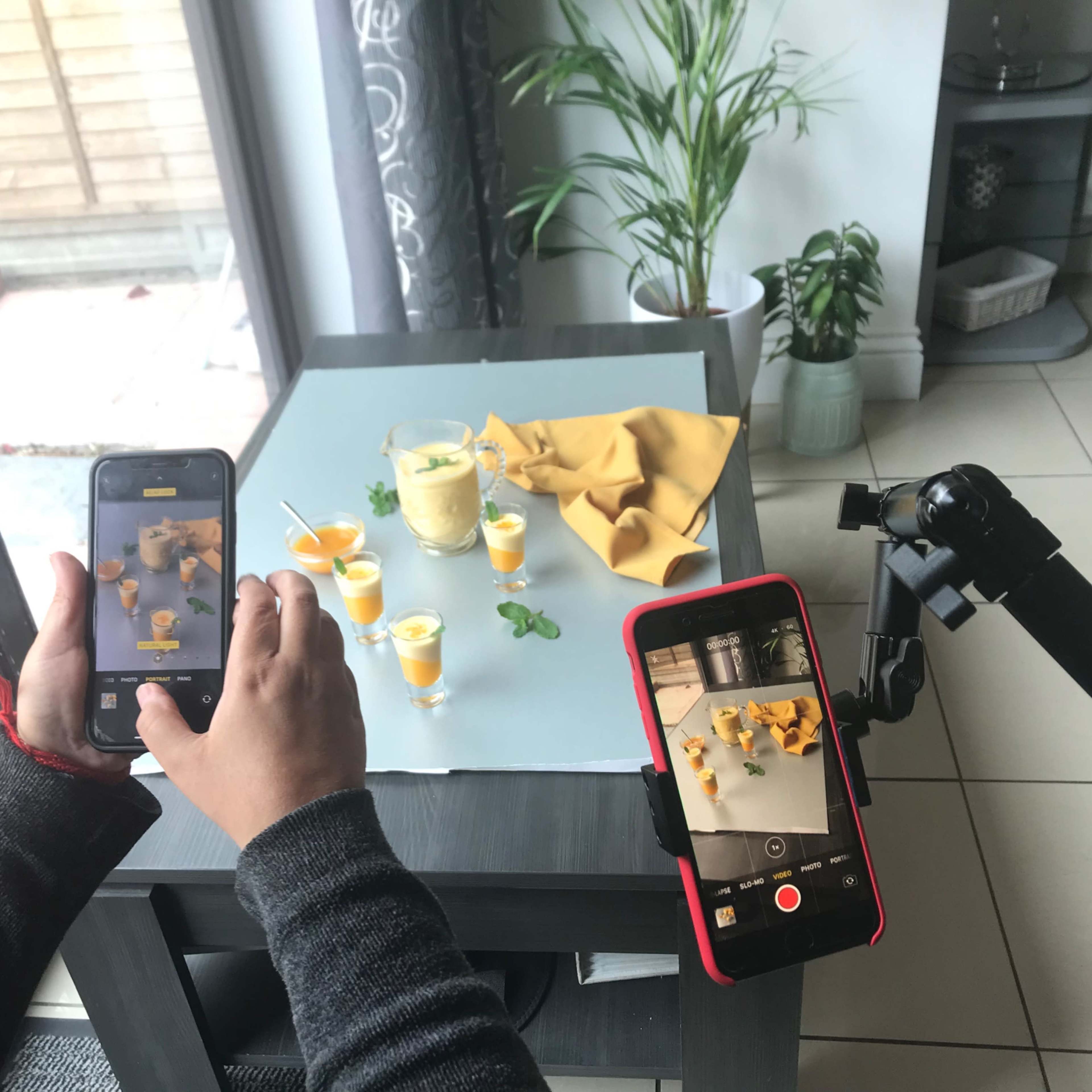 A person is taking photos and videos of small glasses filled with a yellow drink on a table, with a plant in the background.