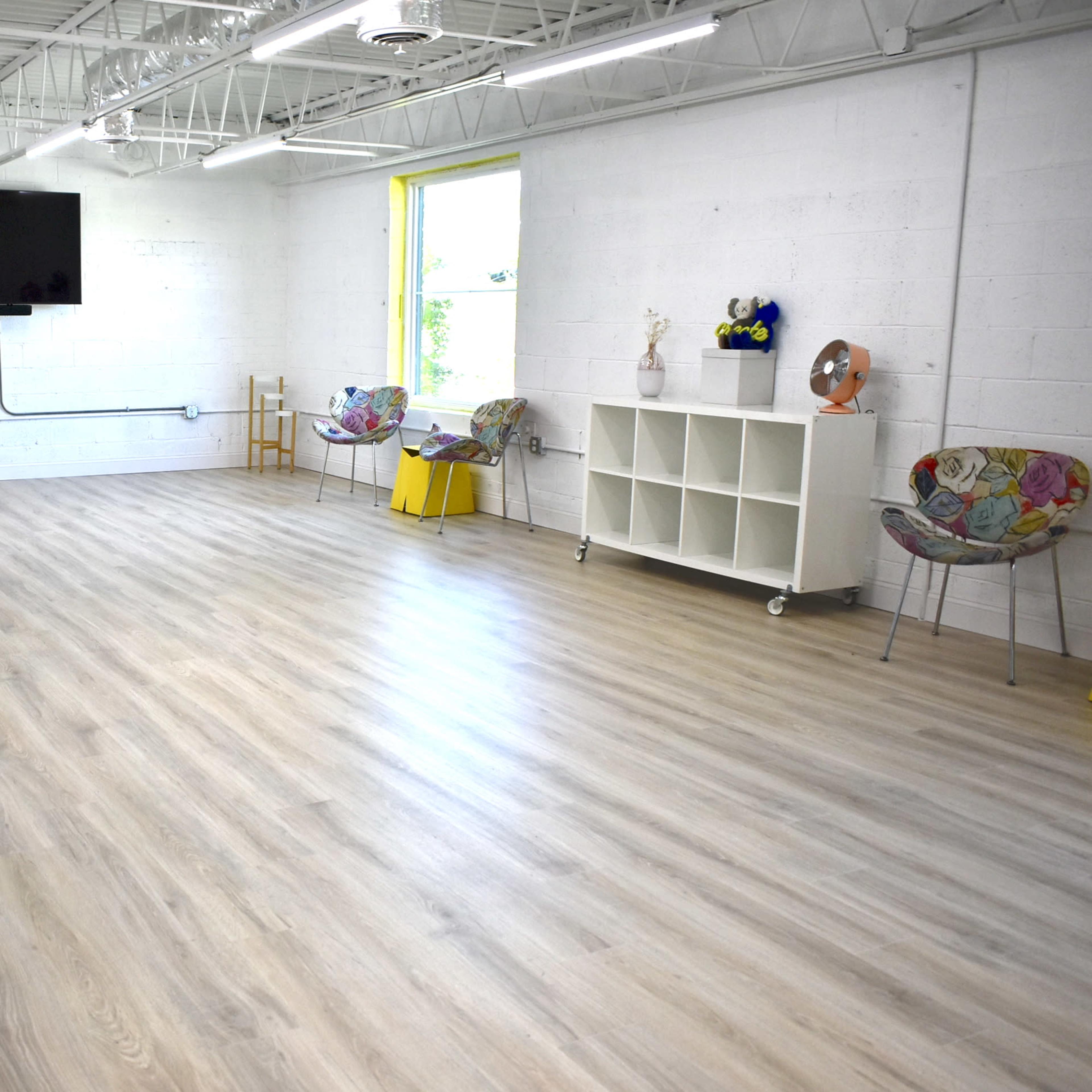 A spacious, well-lit room with light wooden flooring, a wall-mounted television, a window, and various furniture items including chairs and a shelving unit.