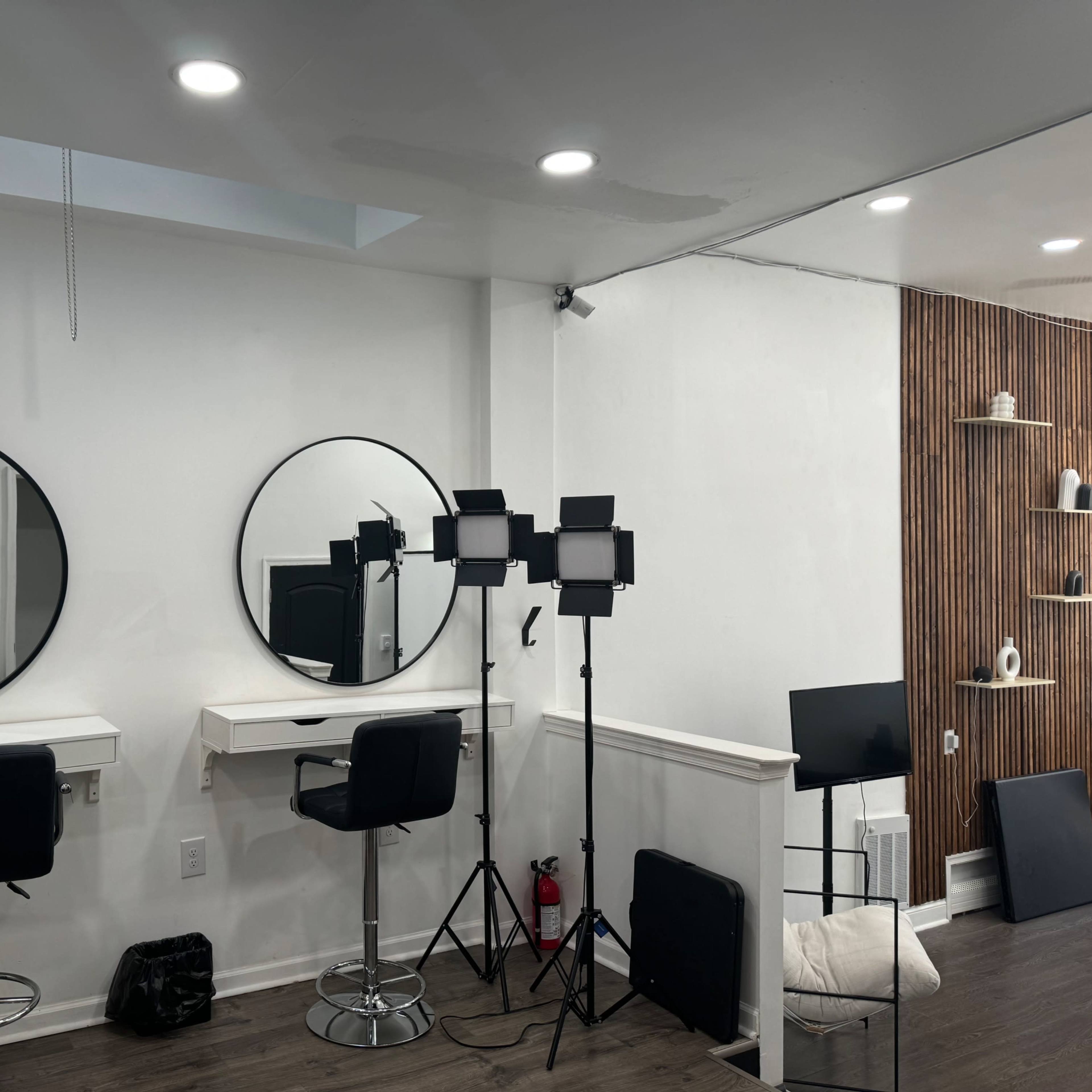 A modern makeup space with two round mirrors, two black bar stools, and studio lighting equipment set up against a white wall.