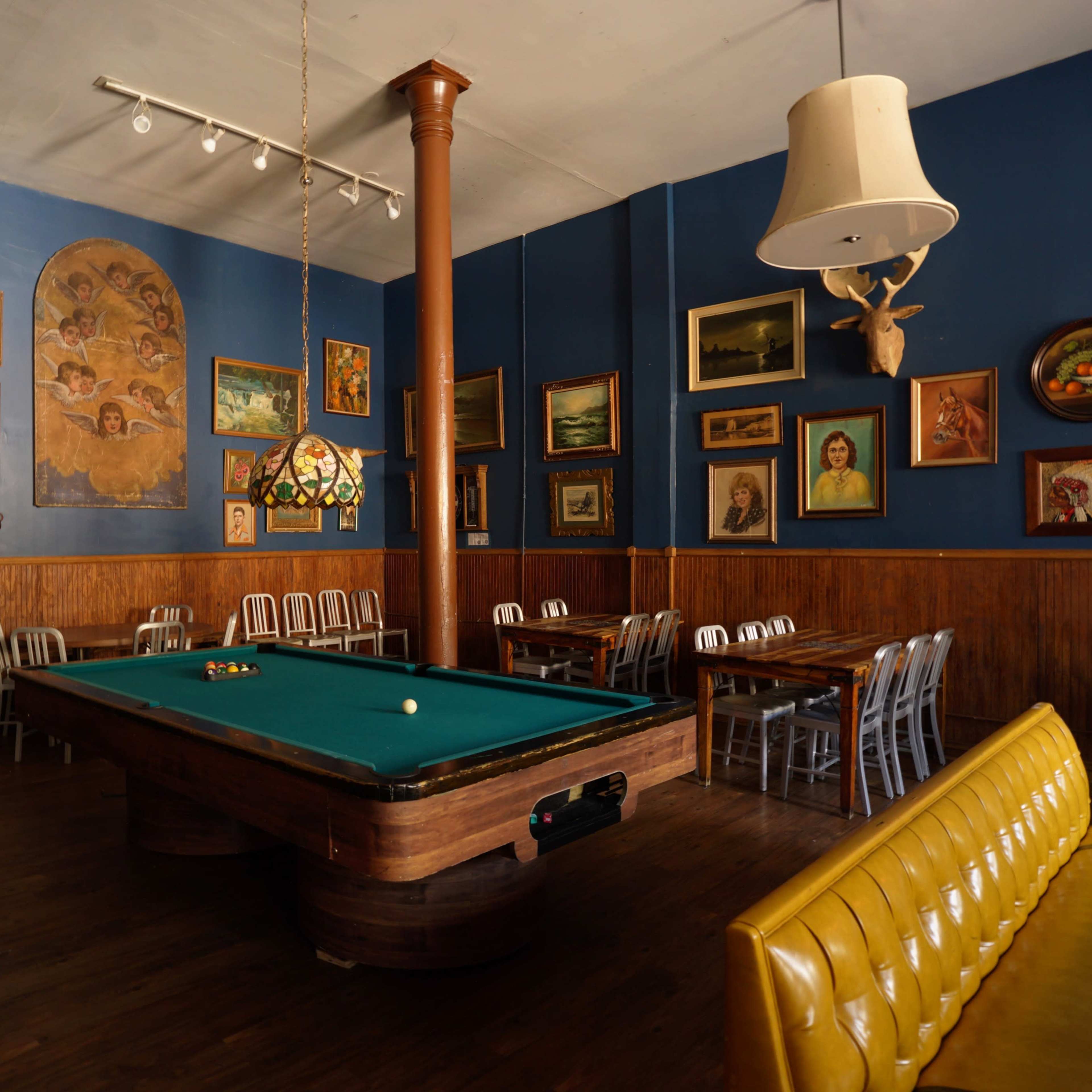 A room featuring a green pool table surrounded by wooden walls adorned with various framed artworks and a mix of seating arrangements.