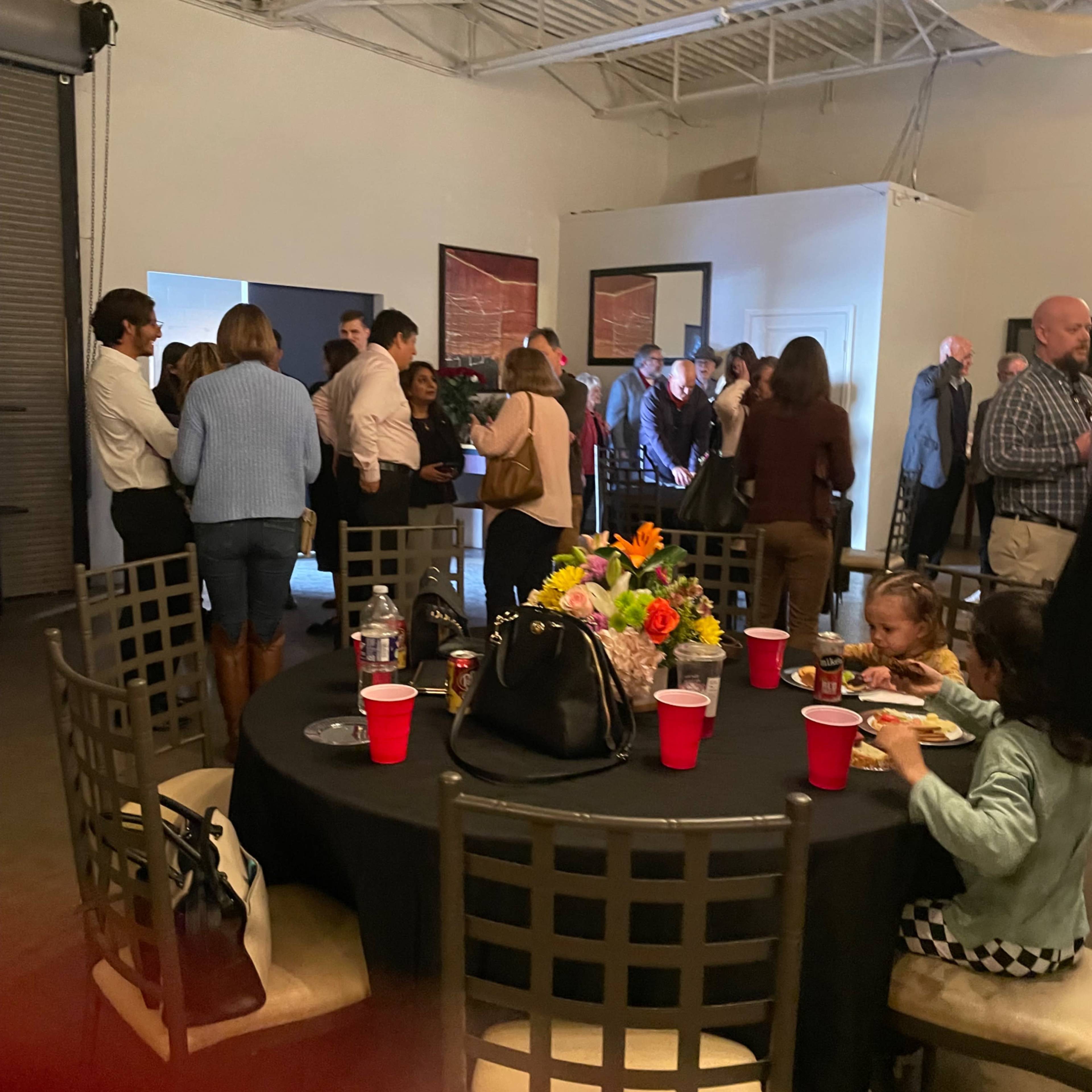 A crowded room filled with people socializing, with a round table in the foreground adorned with flowers and red cups.