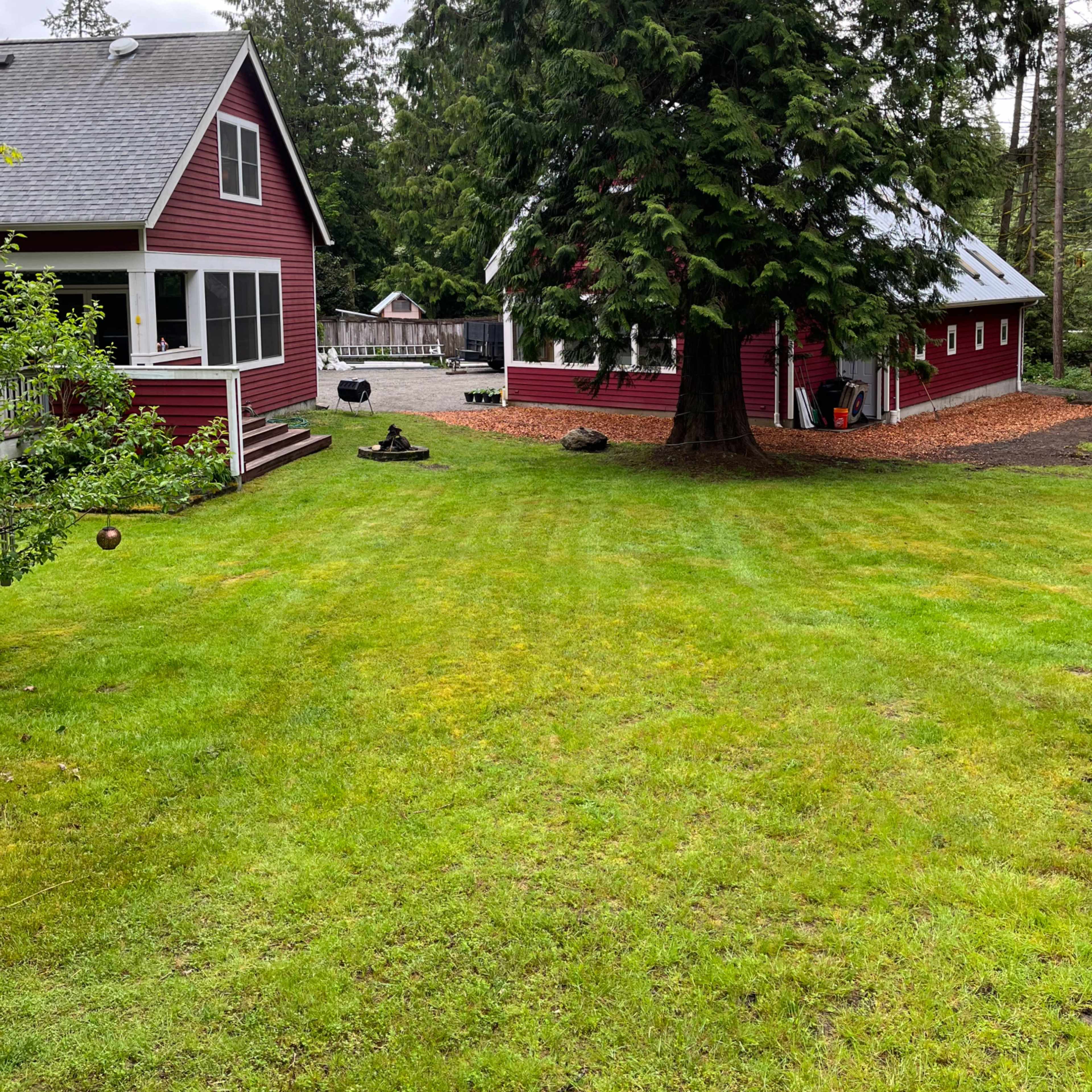 A green lawn surrounded by two red buildings, one a house and the other a barn, with trees in the background.