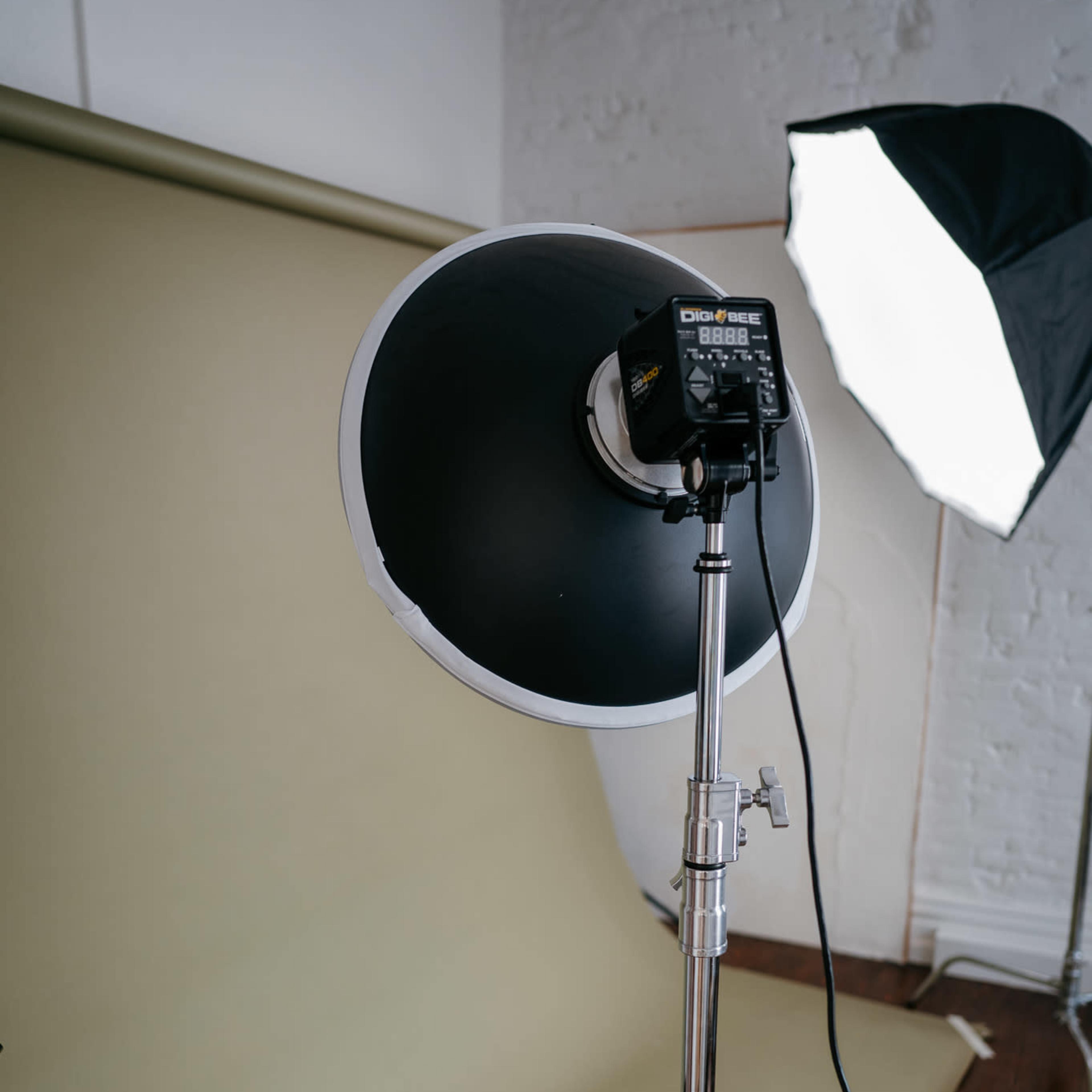 A photography studio setup features a large softbox light and a circular reflector on a light stand in front of a green backdrop.