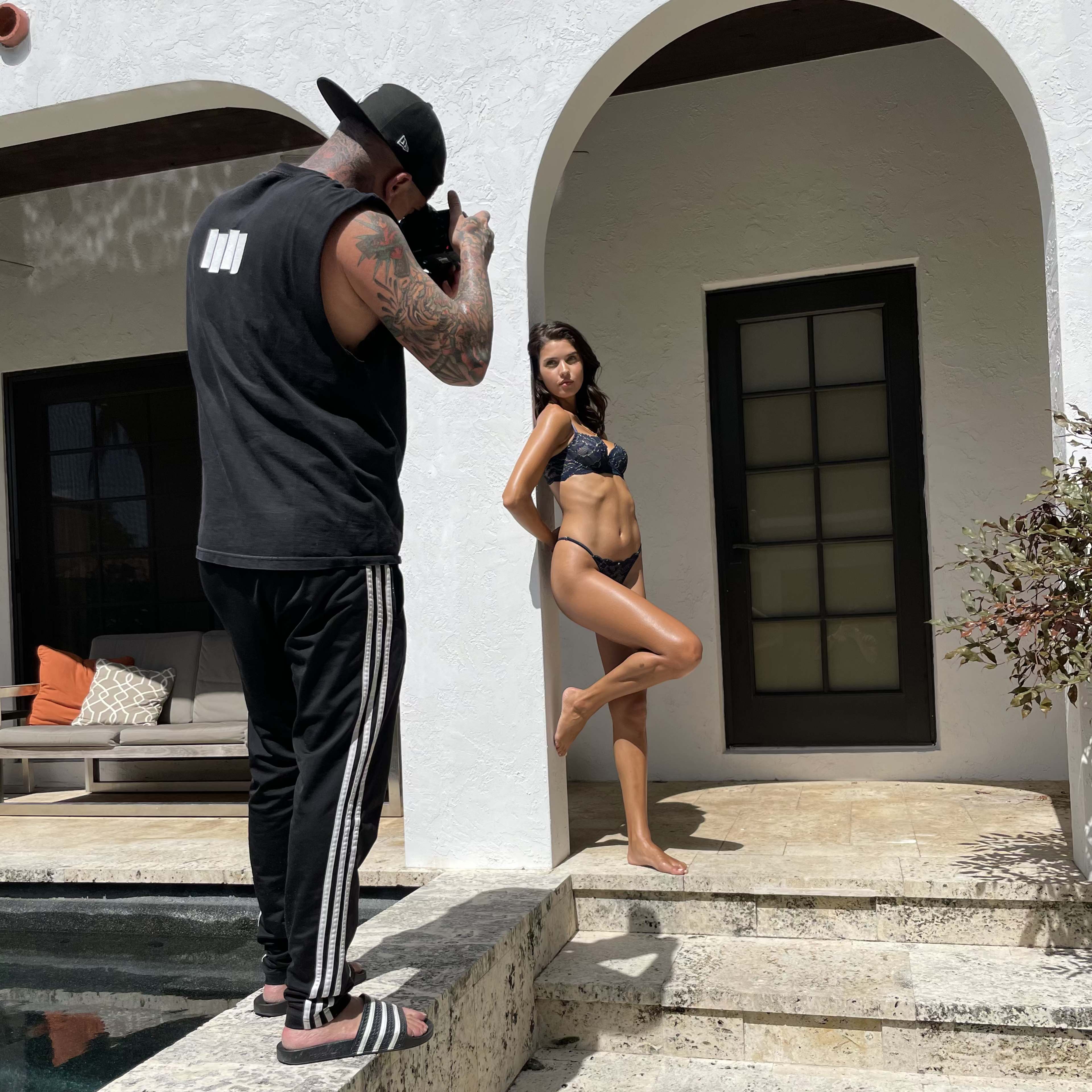 A photographer captures a model in a bikini posing on steps beside a pool at a modern house.