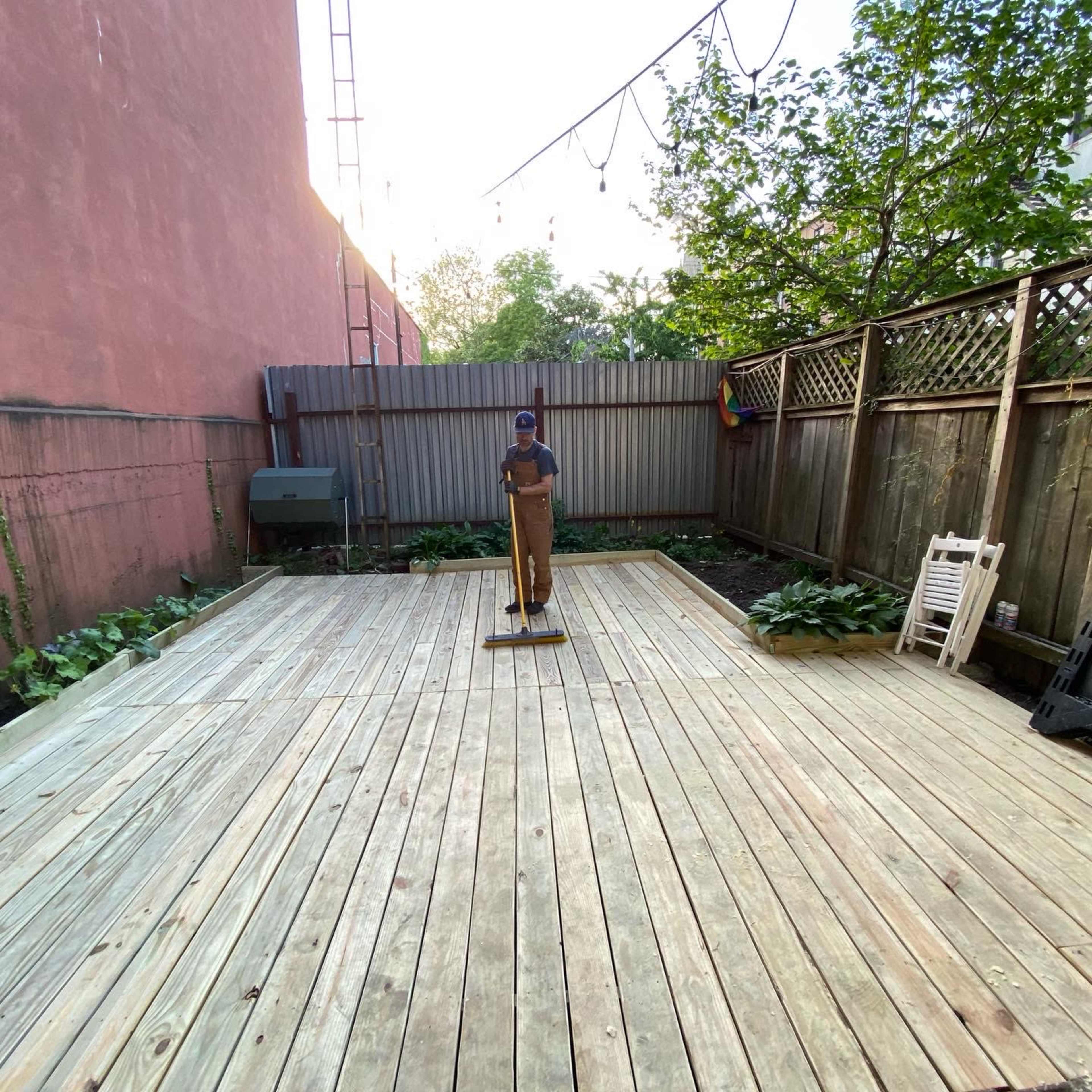 A person in work attire sweeps a wooden deck surrounded by greenery and a wall.