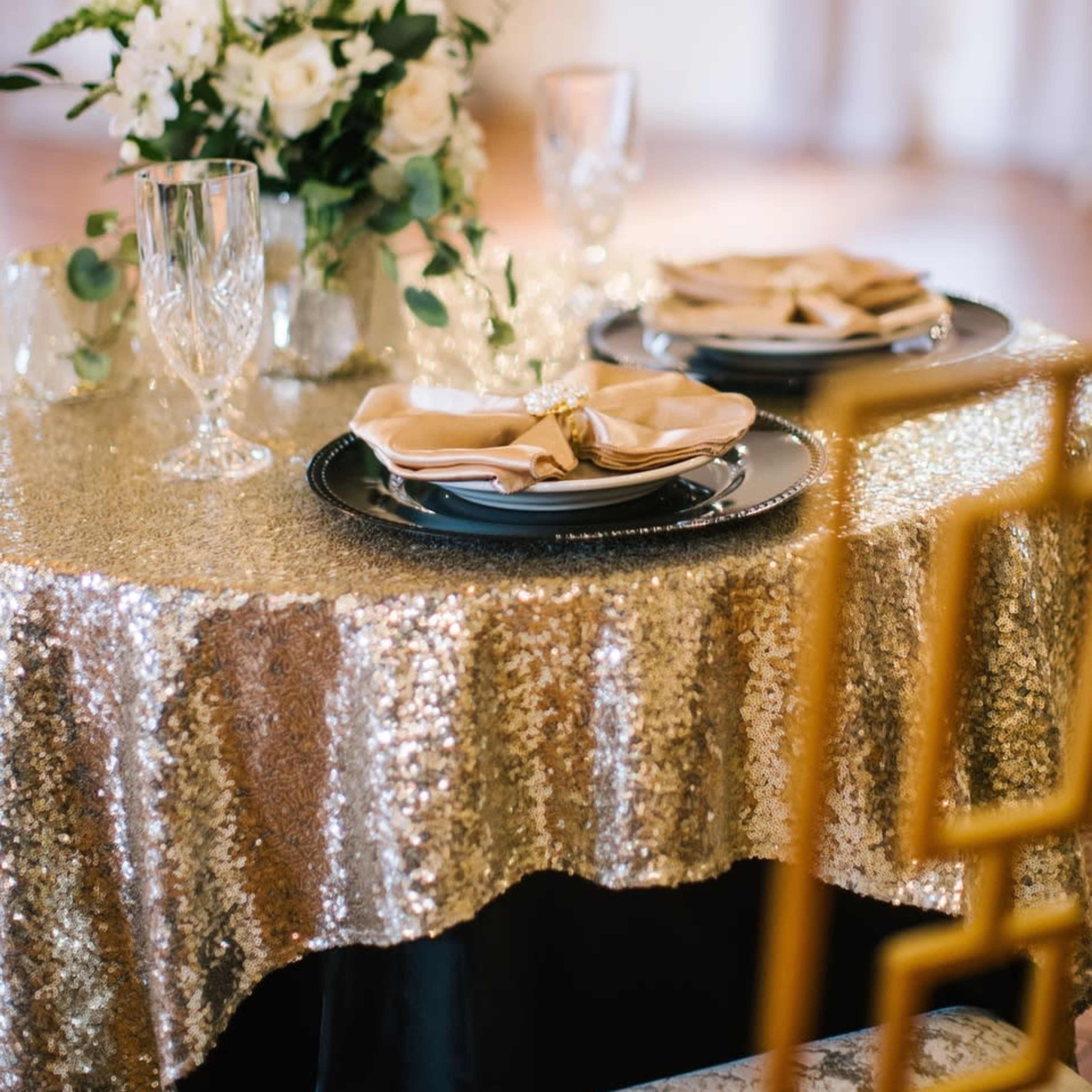 A table is set with elegant plates and napkins, surrounded by decorative glassware and a floral centerpiece, all draped with a sparkling gold tablecloth.