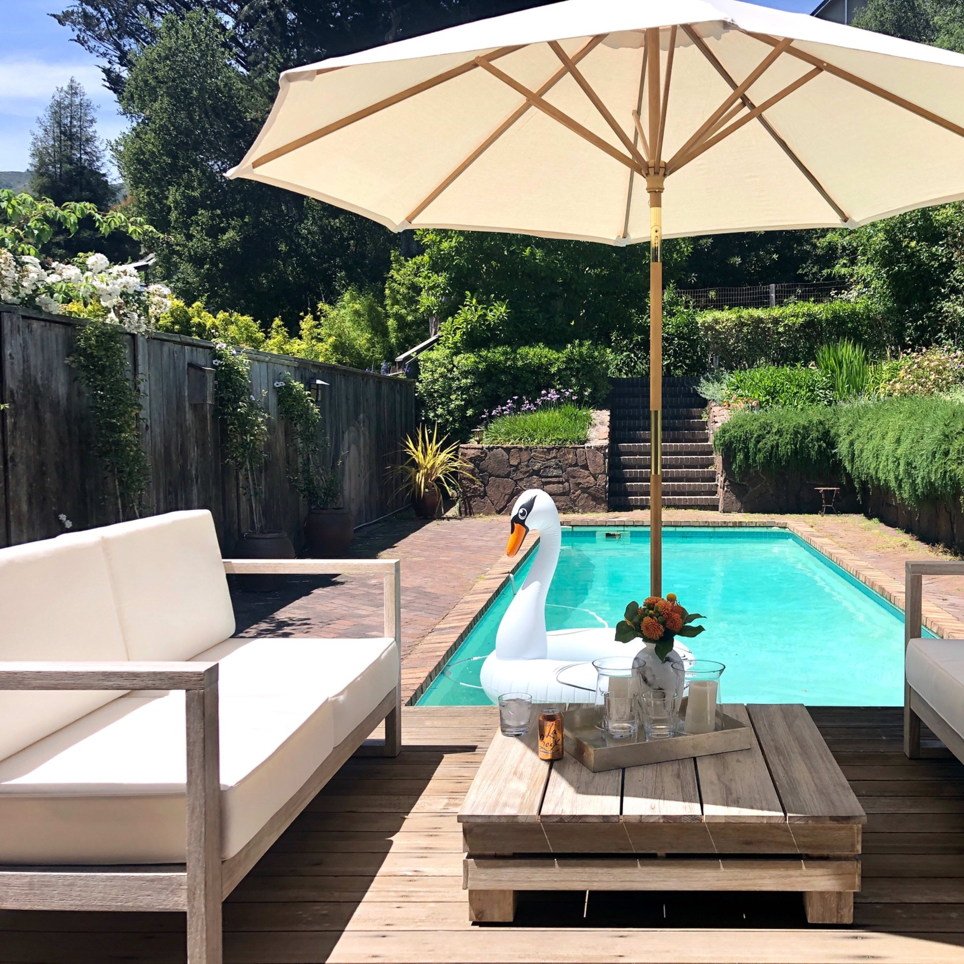 A patio area features two white couches and a wooden table beside a swimming pool, shaded by a large umbrella.