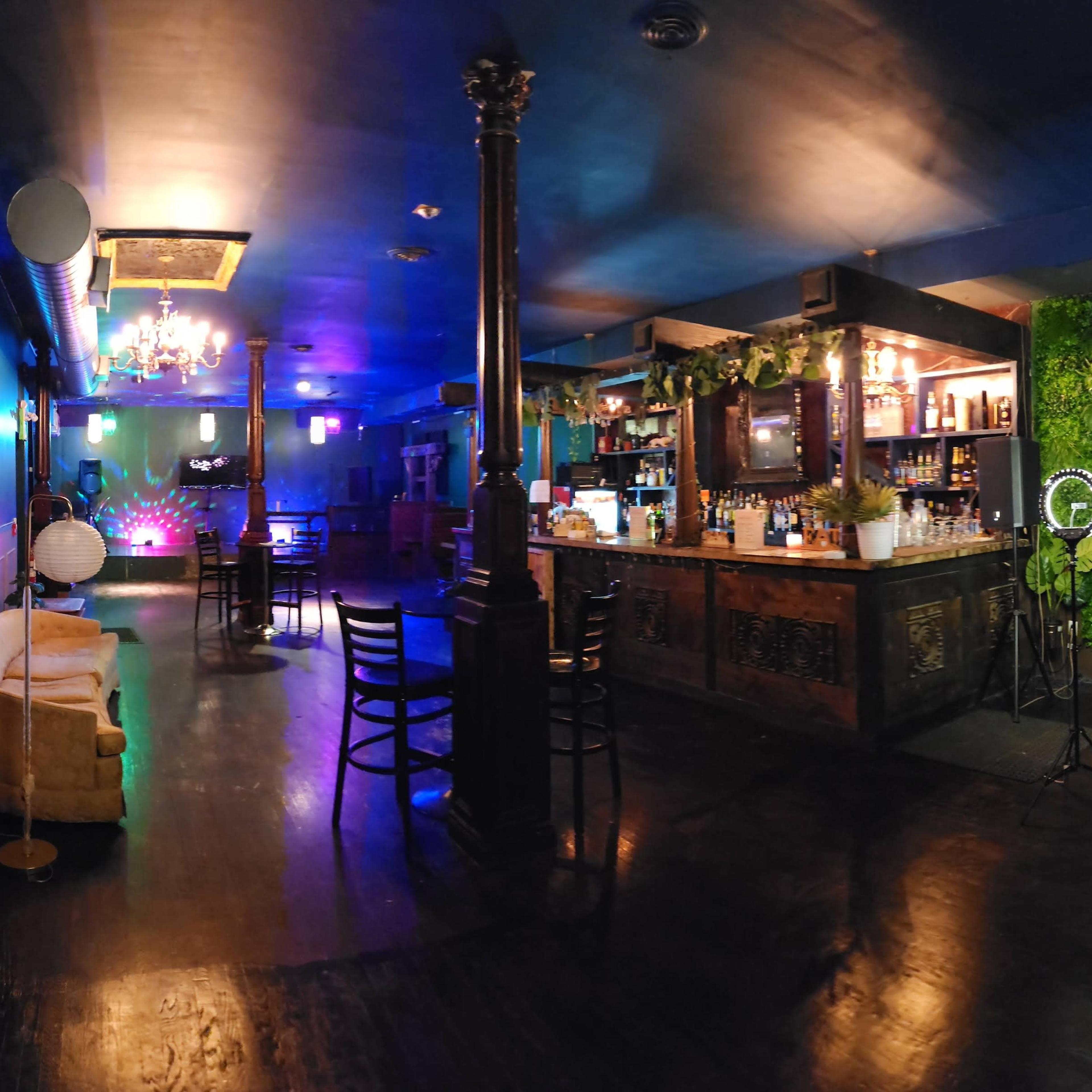 A bar area in a dimly lit lounge with a green plant wall, a wooden bar stocked with bottles, and scattered seating arrangements.