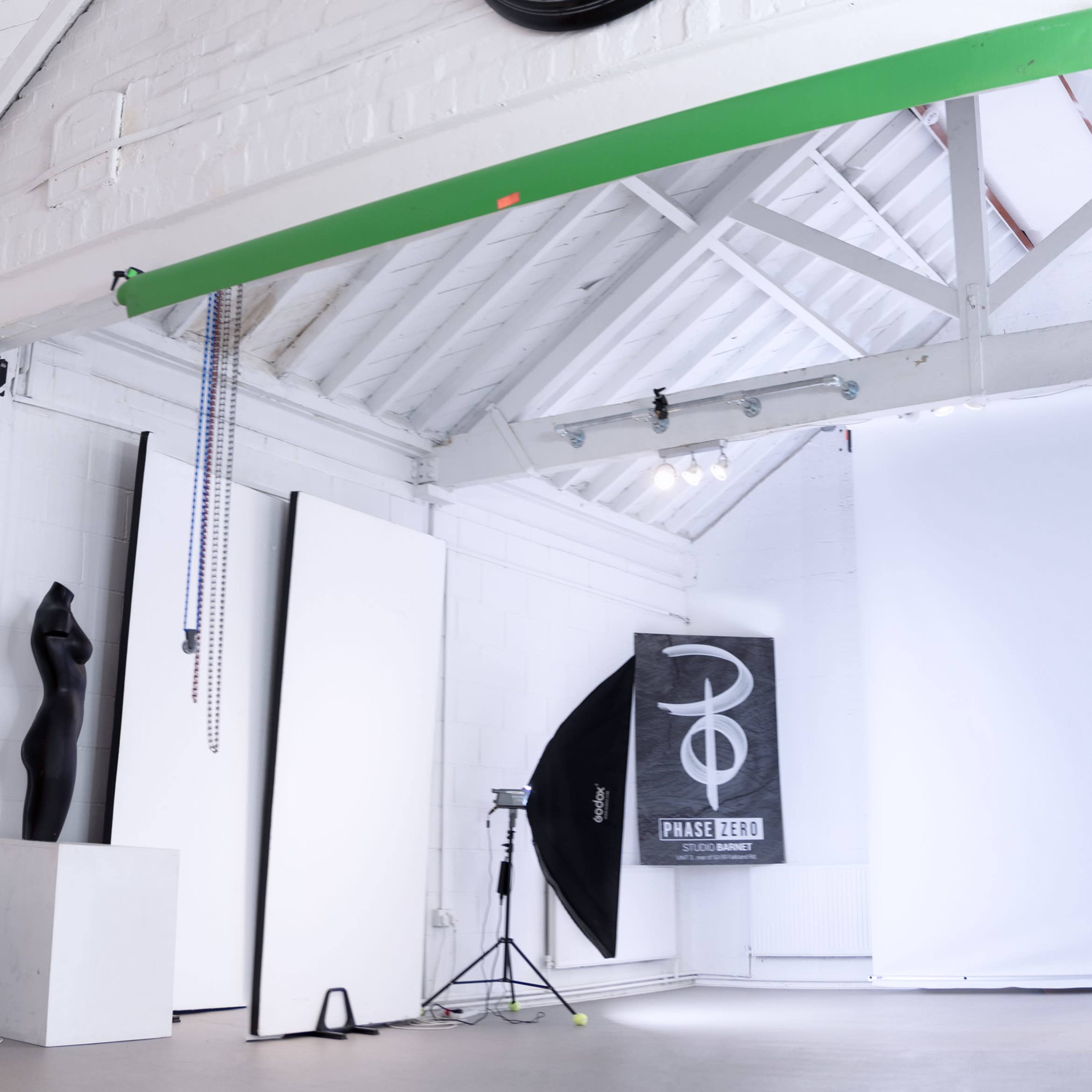 A spacious photography studio with a large clock on the wall, a green beam overhead, and various photography equipment including a softbox and backdrop.