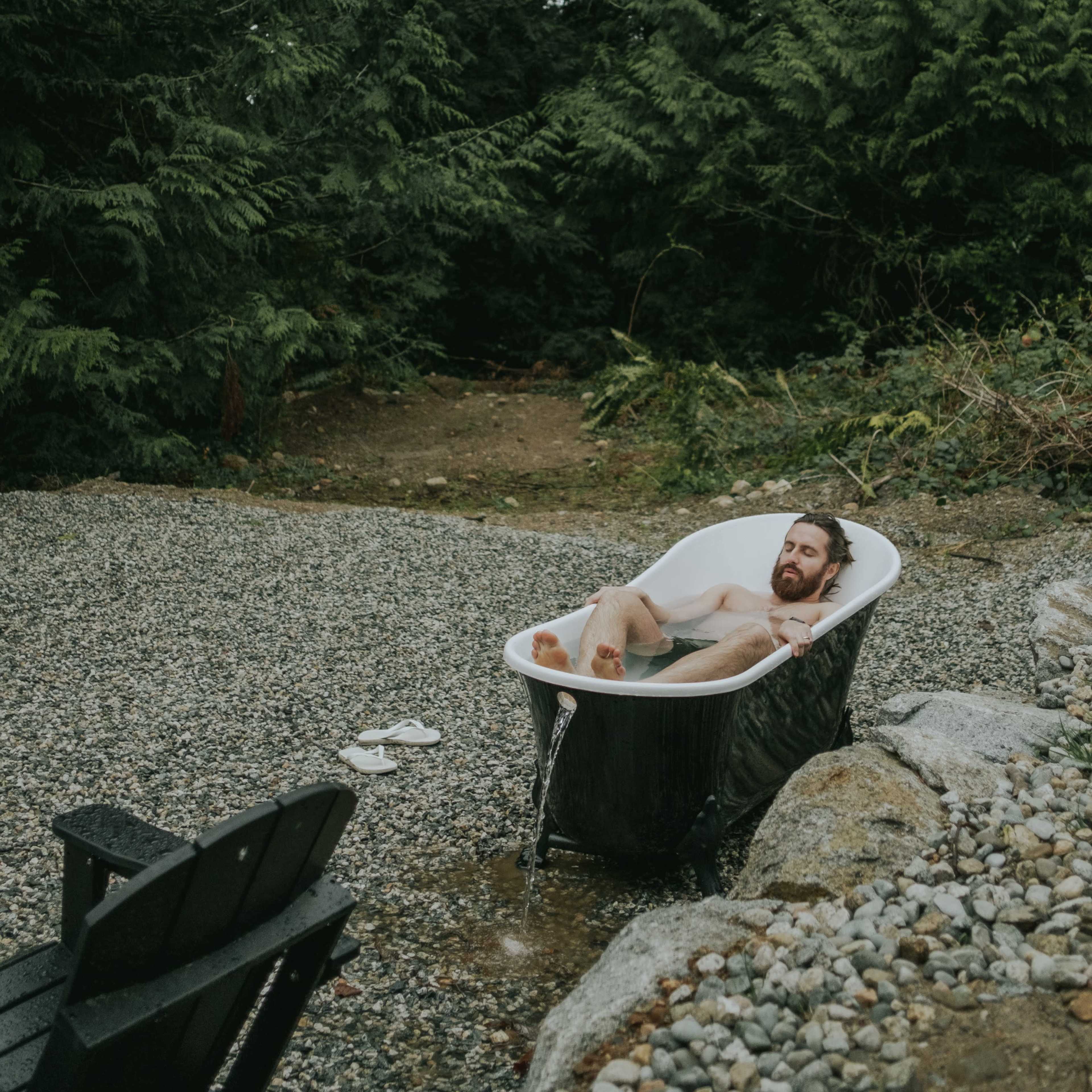 A man is relaxing in a freestanding bathtub surrounded by gravel and greenery in a forested area.