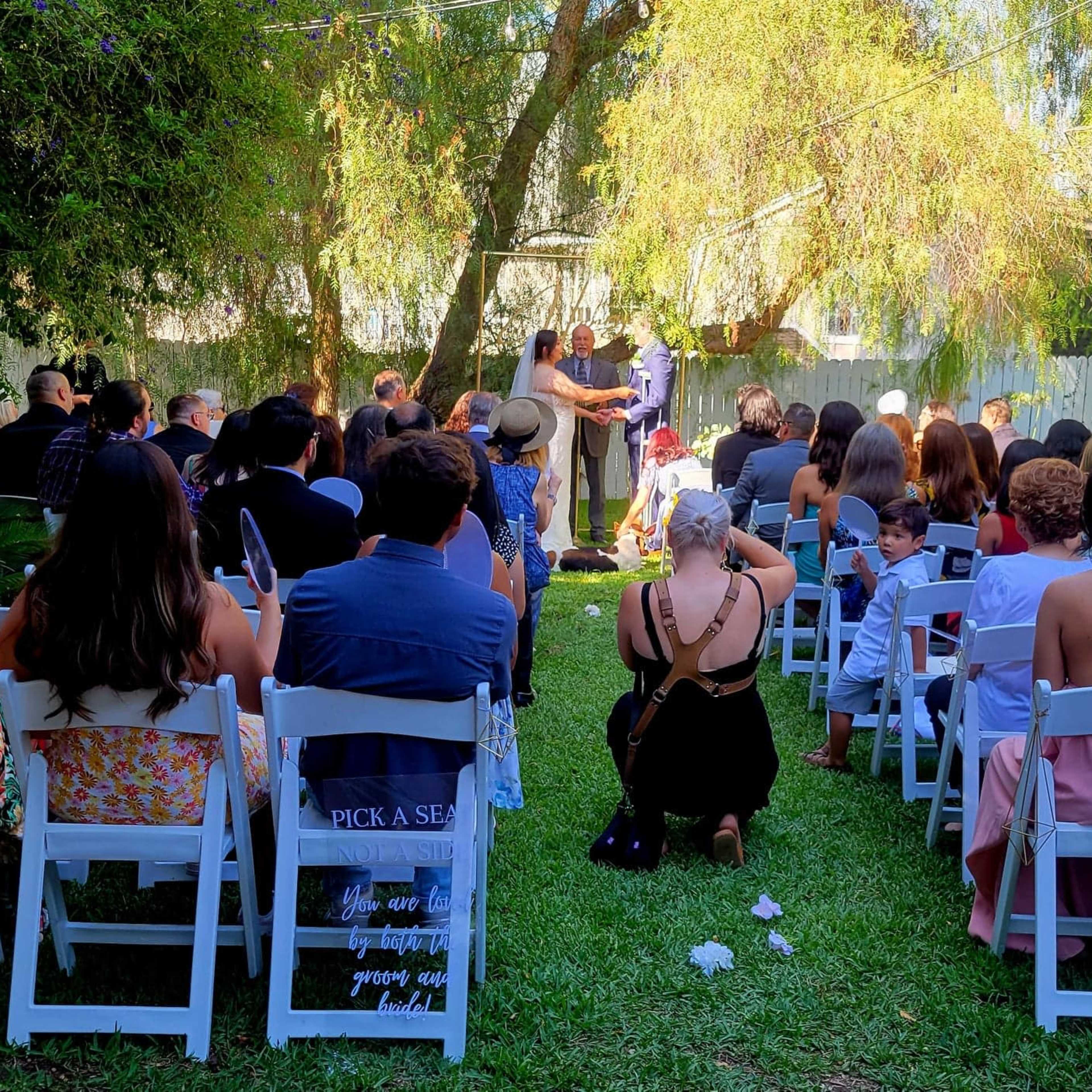 A wedding ceremony is taking place outdoors with guests seated on white chairs in a grassy area.