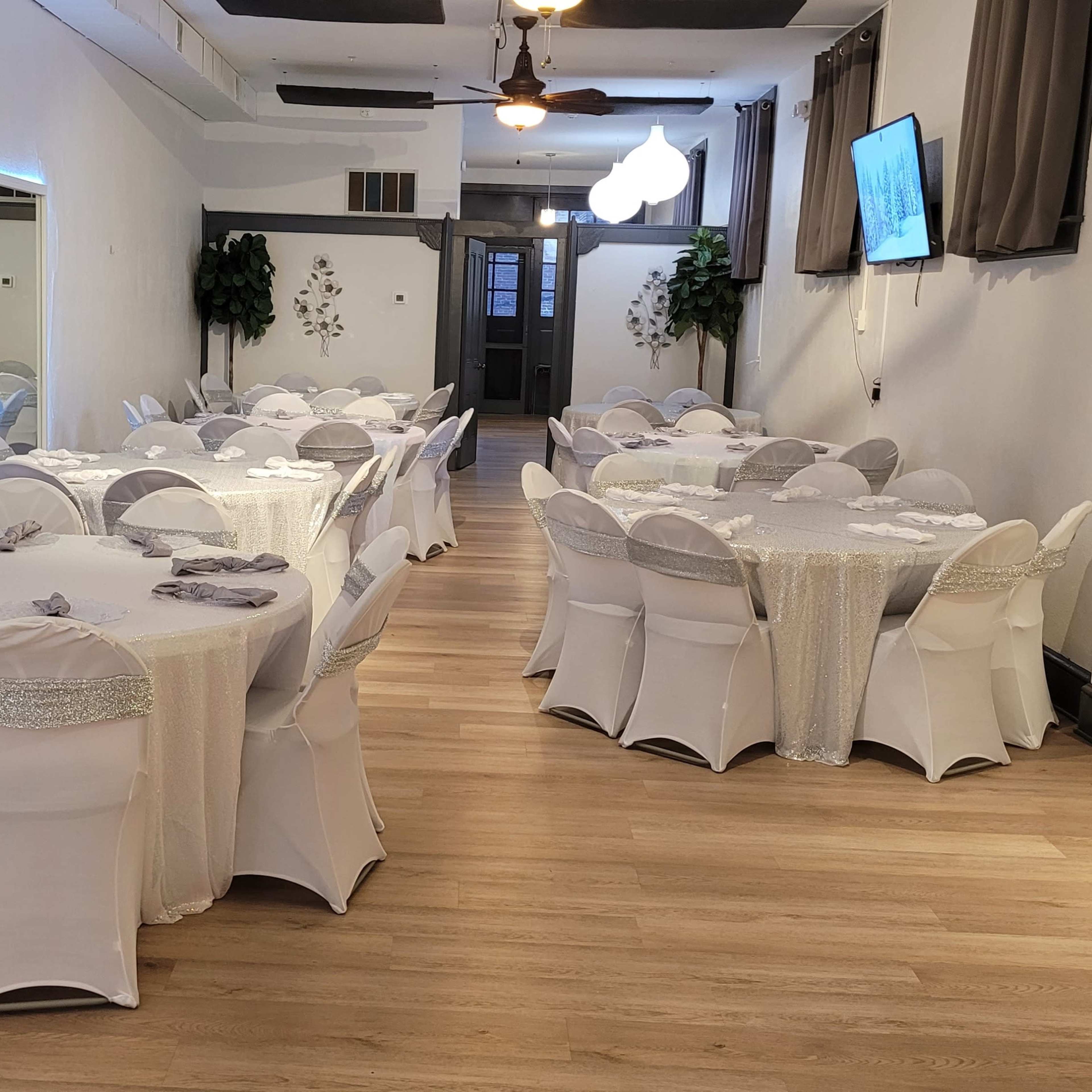 A spacious event room with several round tables dressed in white tablecloths and chairs arranged for a gathering, featuring mirrors and light fixtures on the walls.