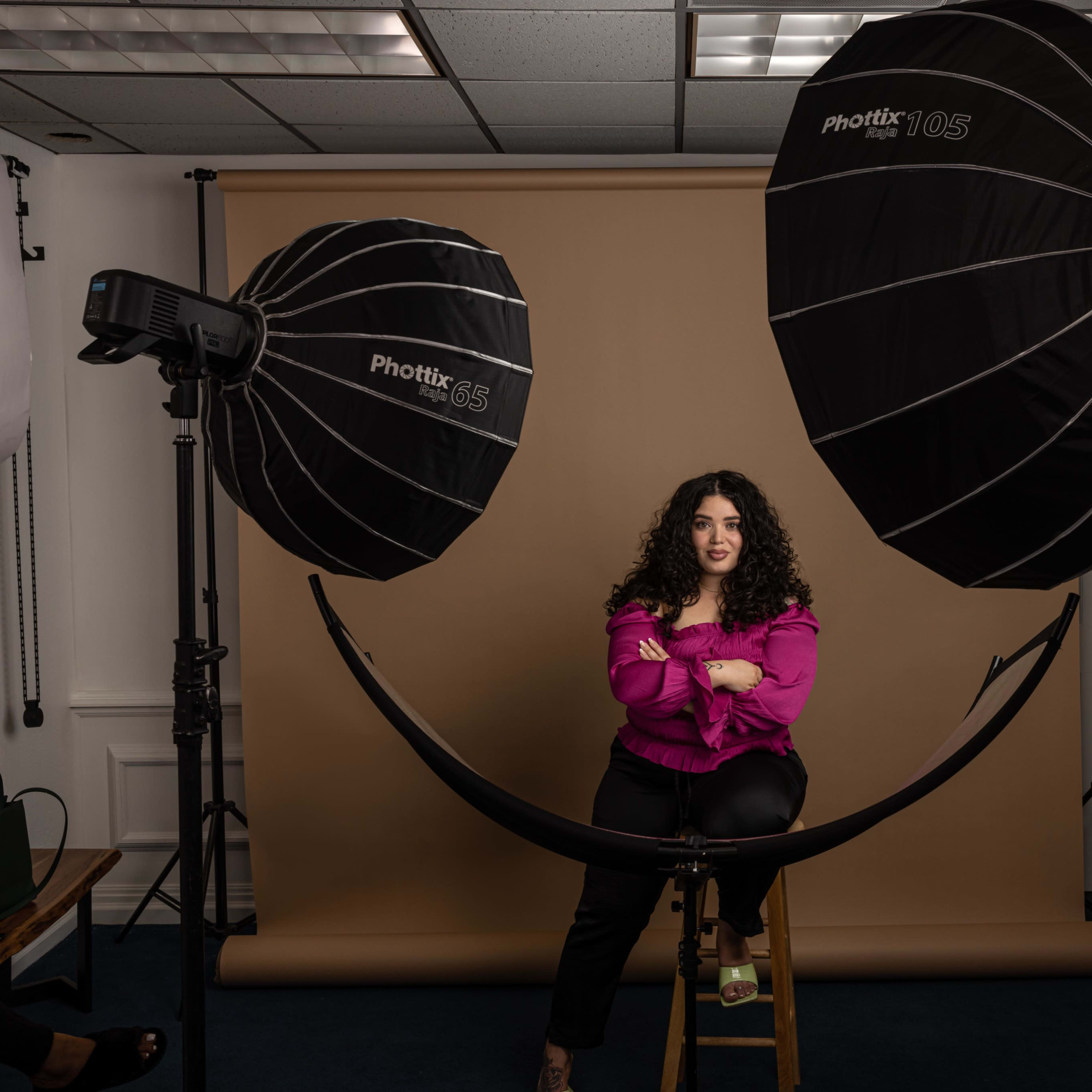 A person sits on a stool in front of a beige backdrop, illuminated by two large softbox lights on either side.