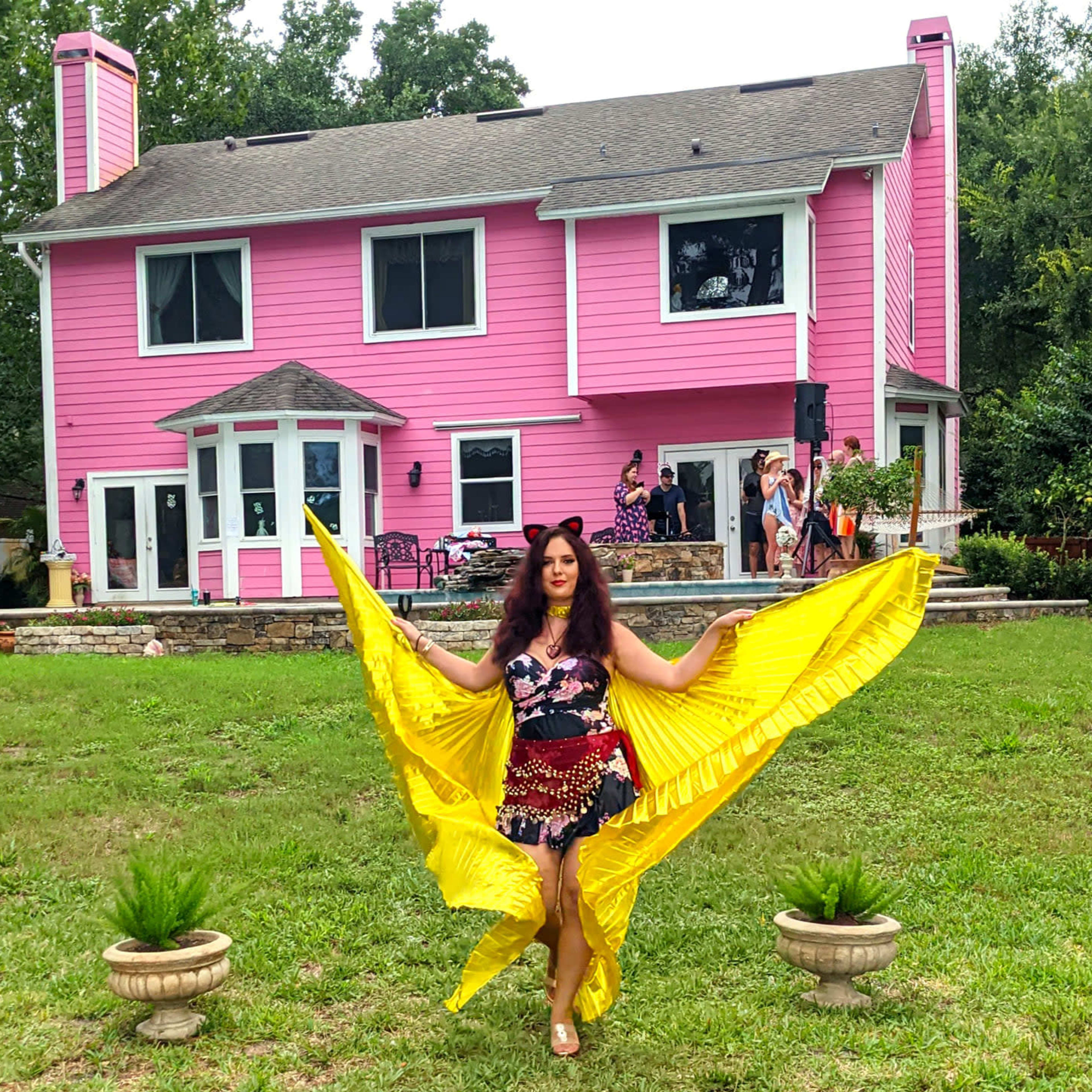 A woman in a colorful outfit holds a yellow fabric as she stands in front of a large pink house surrounded by greenery.