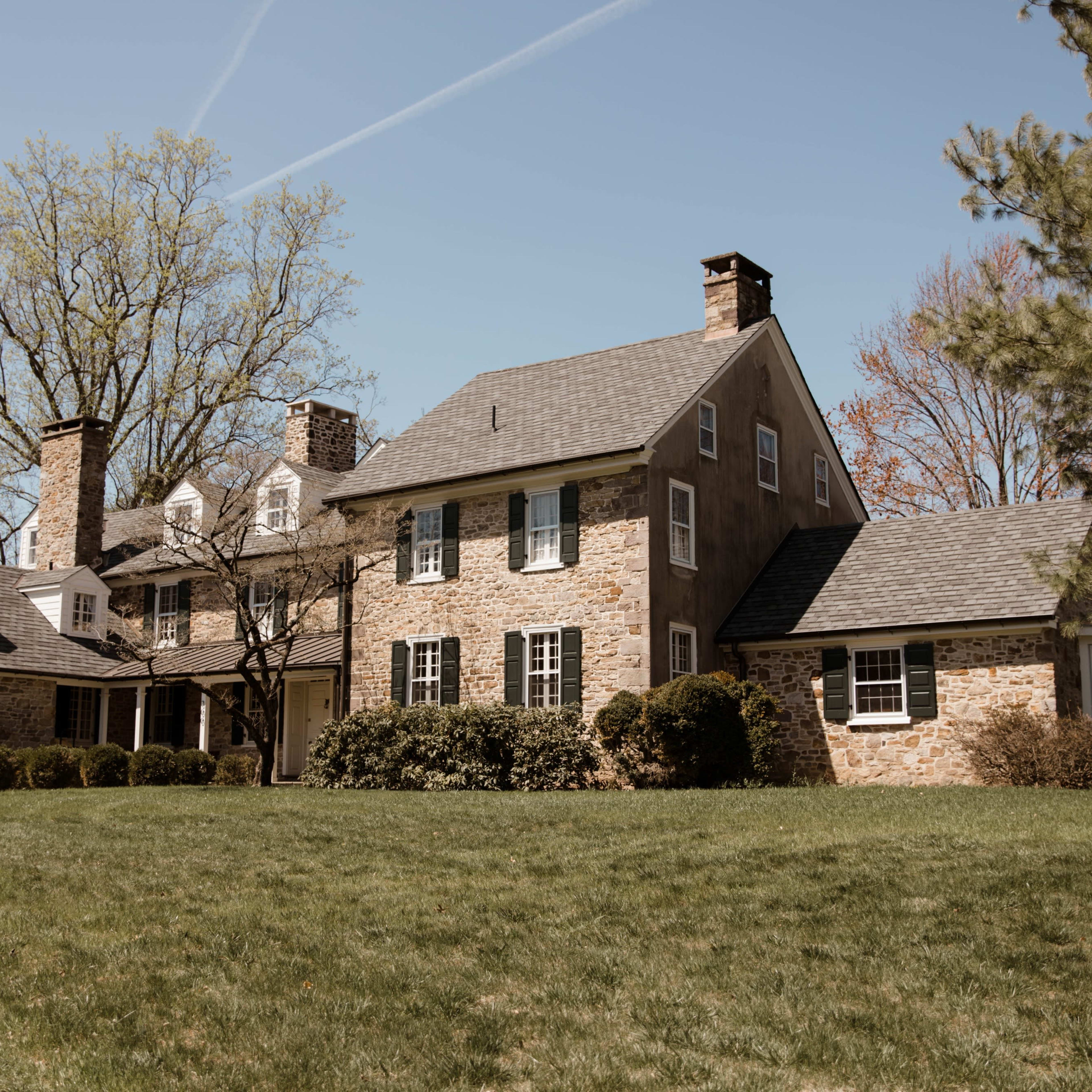 A large stone house with multiple chimneys is surrounded by green grass and trees on a clear day.
