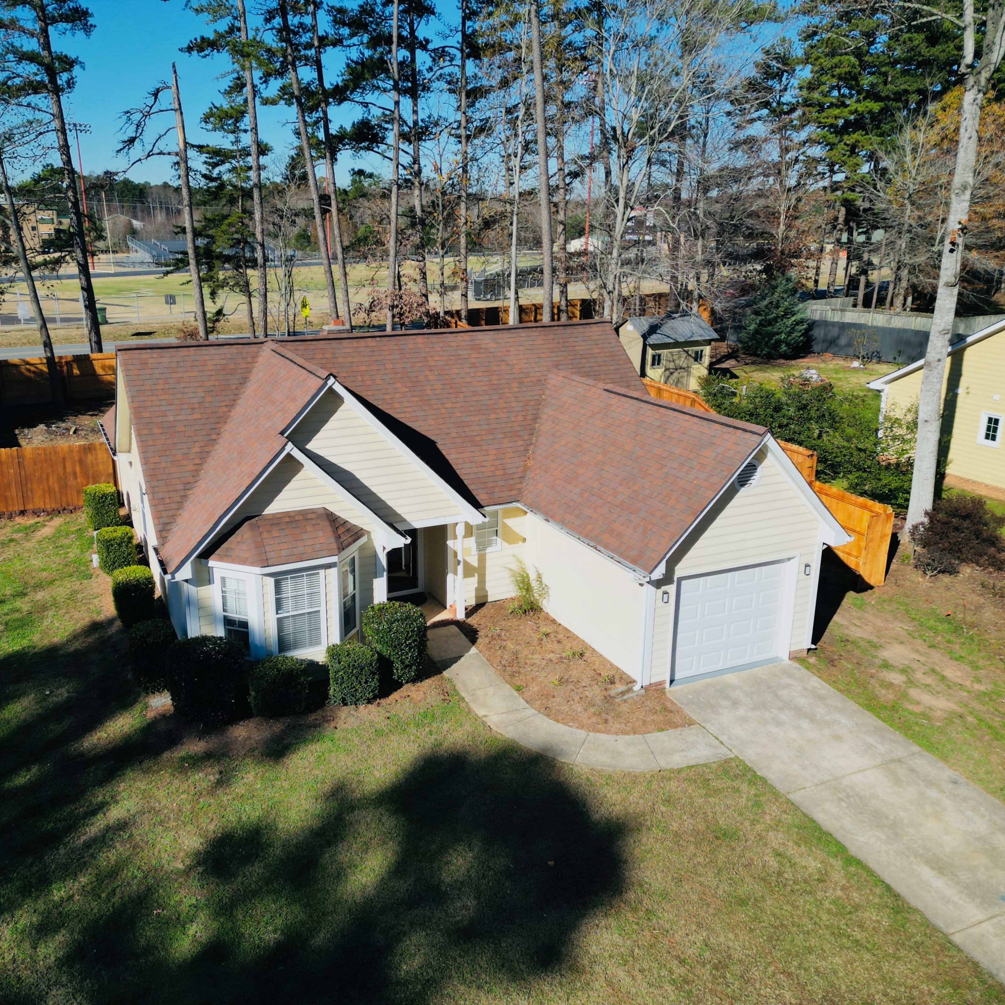A single-story house with a brown roof and a neatly landscaped yard is located in a suburban neighborhood, surrounded by trees and a wooden fence.