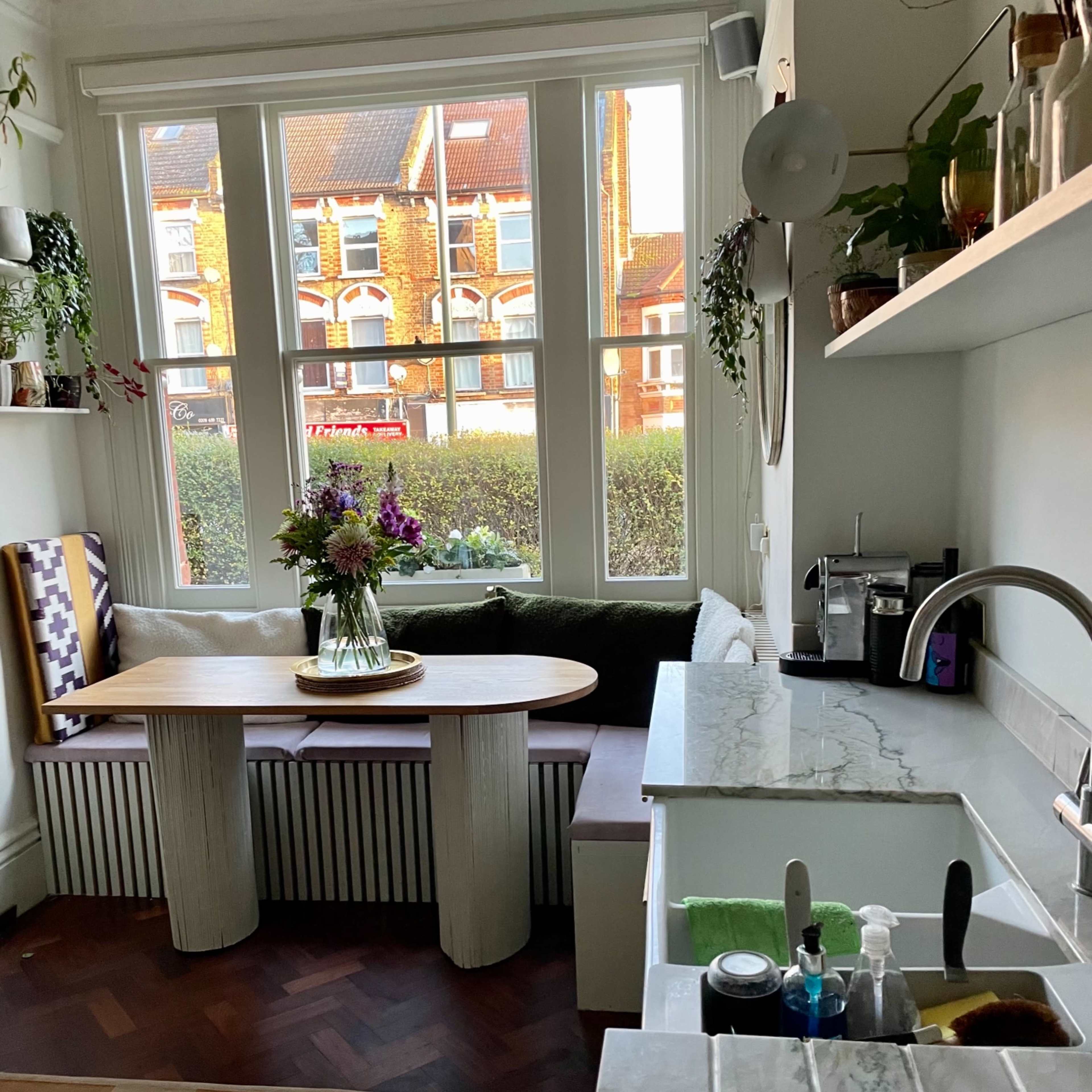 A well-lit kitchen area with a wooden table and a dark sofa, framed by large windows overlooking an outdoor garden.