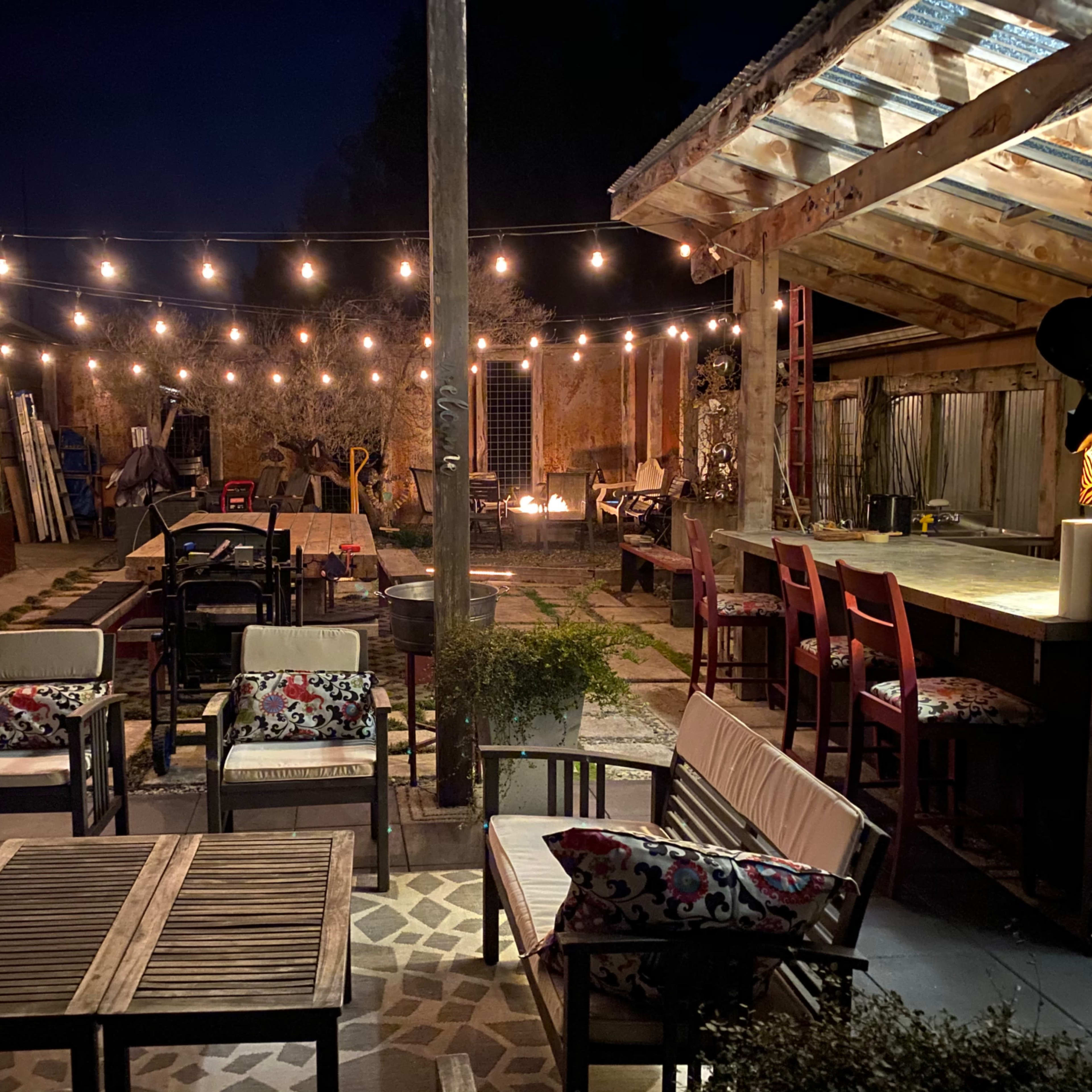 A nighttime outdoor patio area with string lights overhead, wooden seating, and a fire pit in the corner.