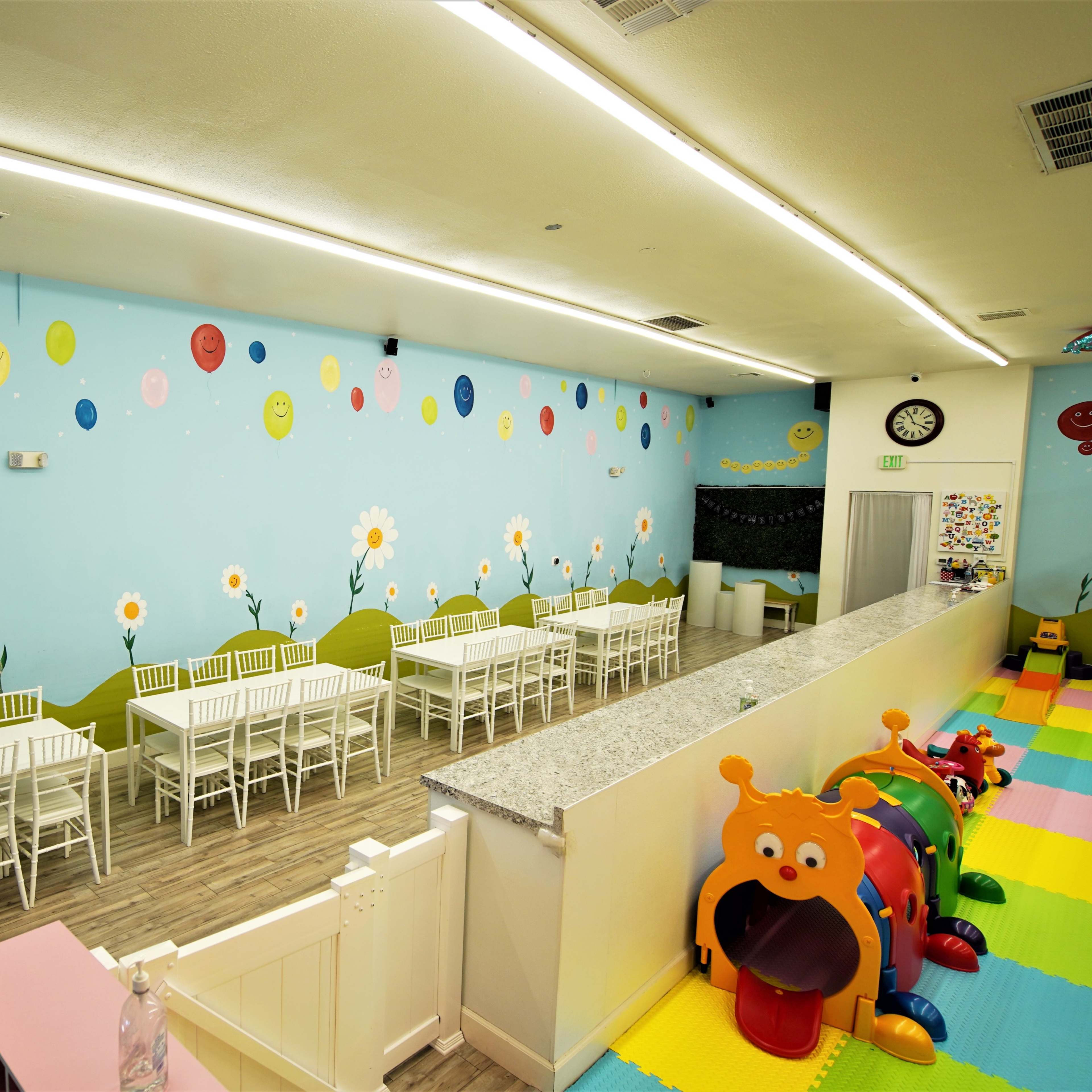 A brightly colored indoor playroom with decorated walls, featuring tables and chairs arranged neatly along one side and play equipment in one corner.