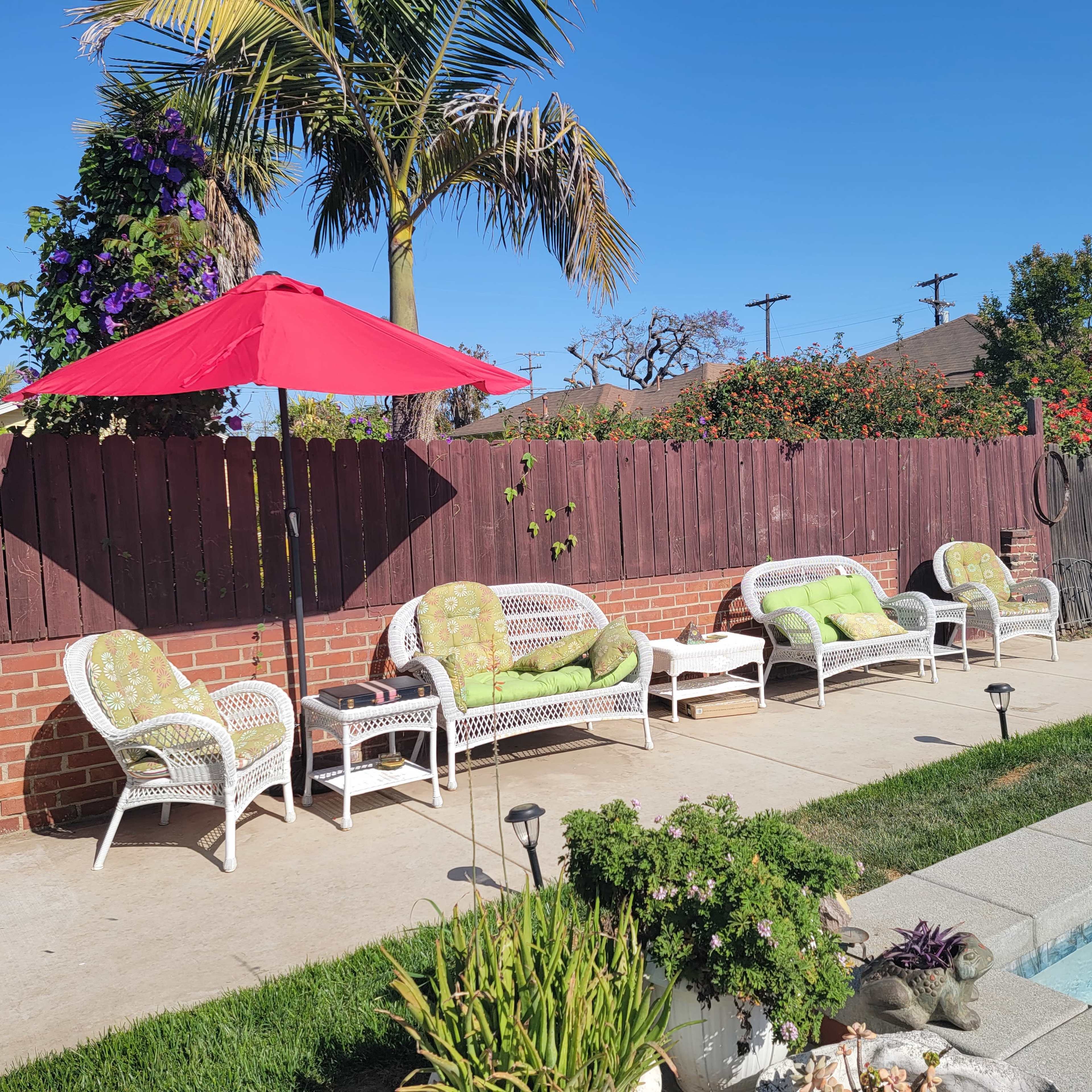 A patio area features white wicker furniture with green cushions, a red umbrella, and a well-maintained swimming pool surrounded by landscaping.