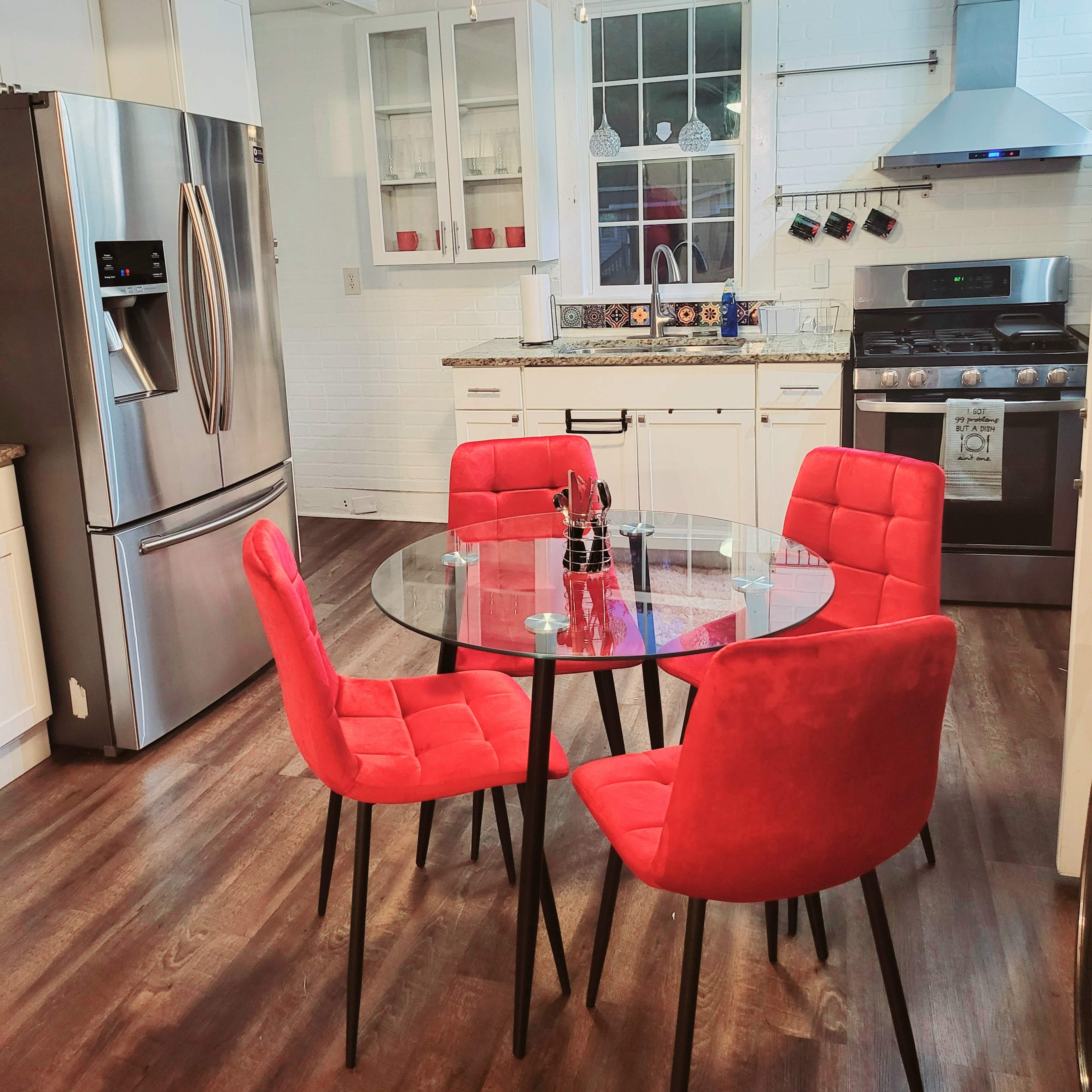 A modern kitchen features a round glass dining table with four red chairs, stainless steel appliances, and a white backsplash.