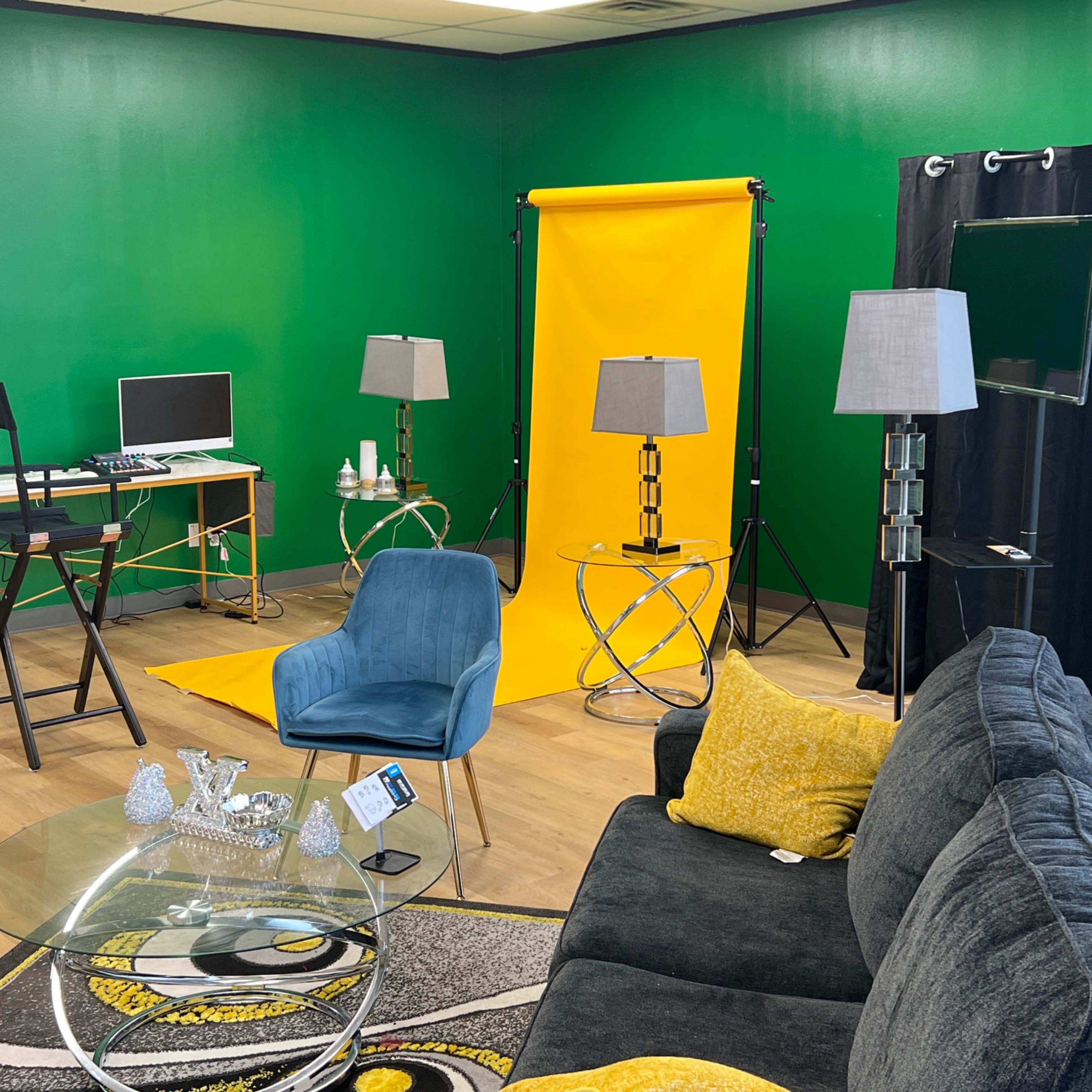 A studio setup featuring a green wall, yellow backdrop, a desktop computer, lighting equipment, a gray sofa, and a glass coffee table.
