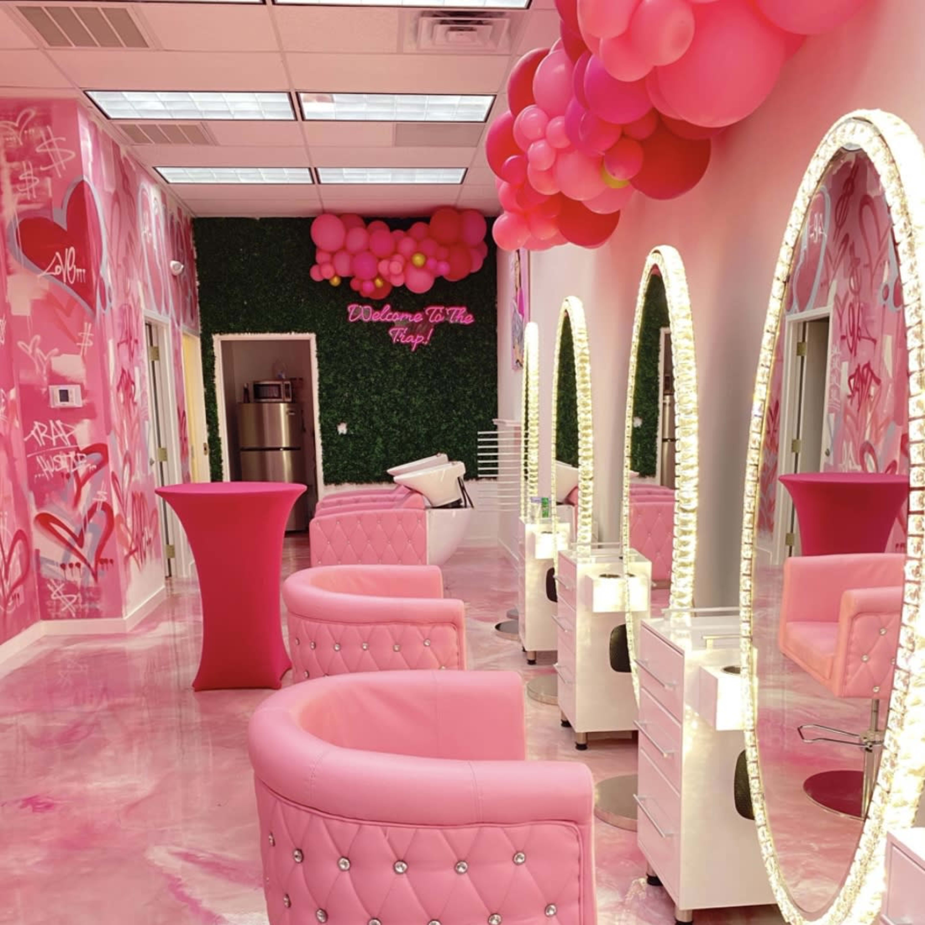 A brightly lit salon with pink furniture, decorative wall art, and modern styling stations arranged along a glossy floor.