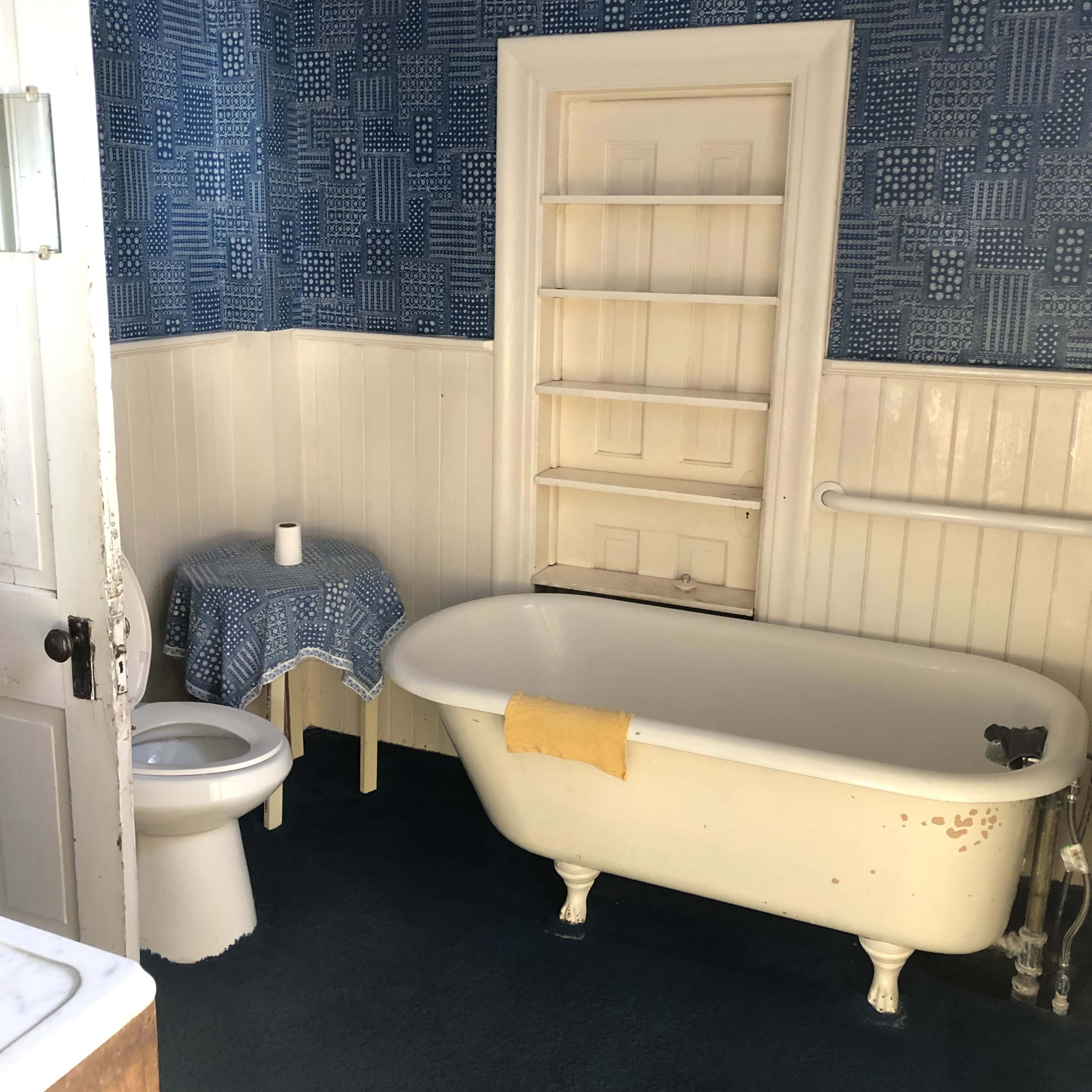 A bathroom with a vintage clawfoot bathtub, a toilet, and a small table with a candle, all set against blue patterned wallpaper.