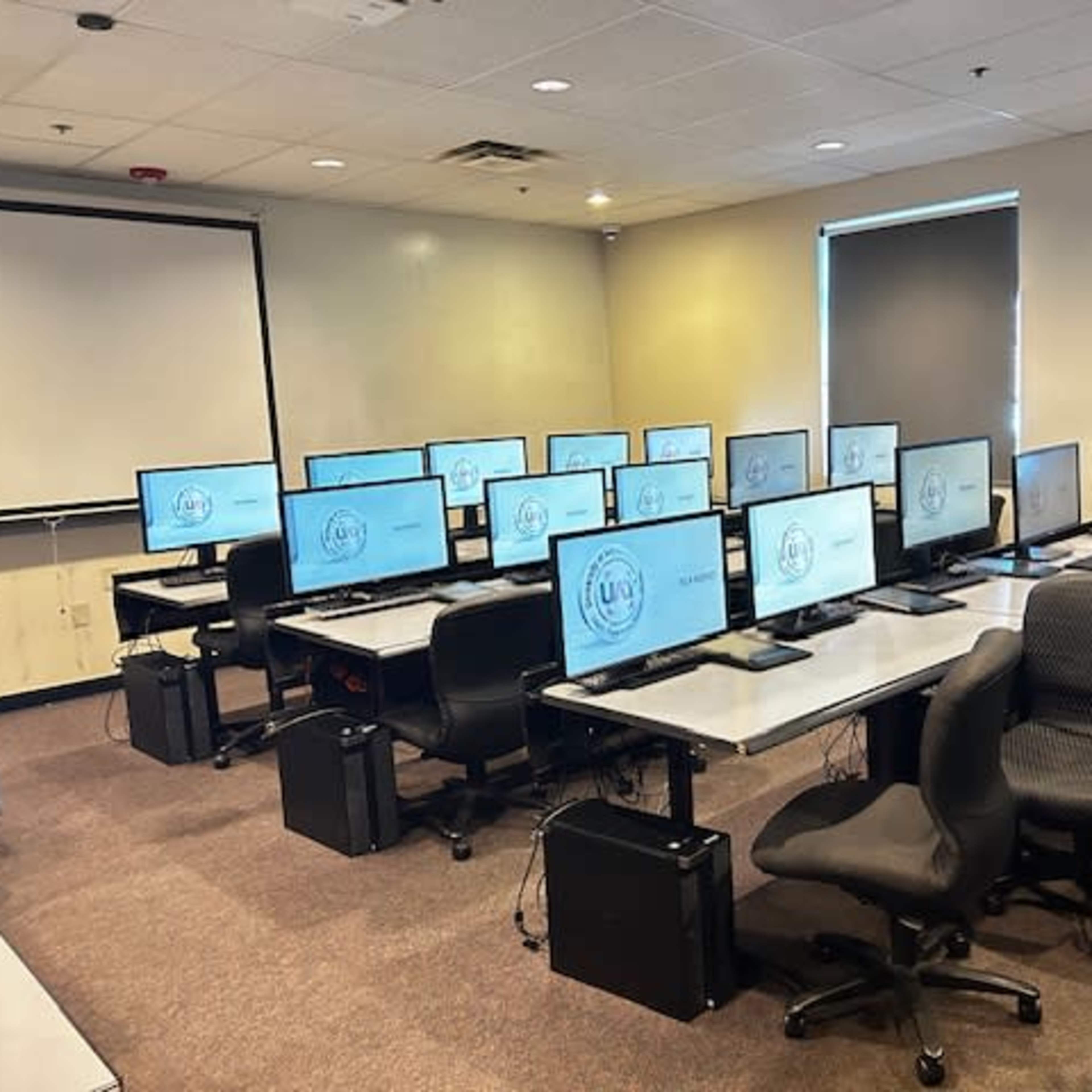 A classroom is arranged with multiple computer stations, each featuring a monitor and chair, facing a presentation screen.