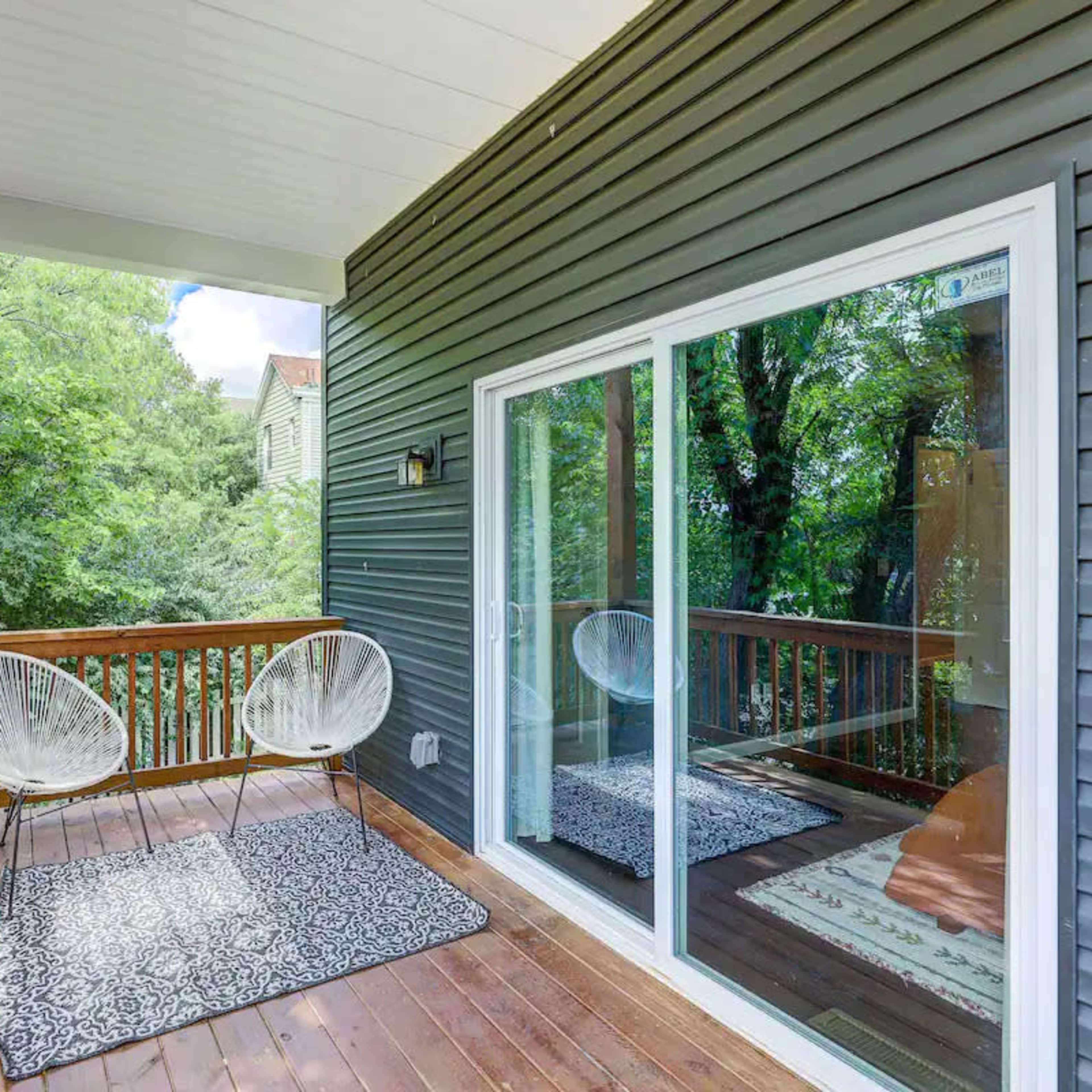A wooden deck with two white chairs and a patterned rug, surrounded by green trees.