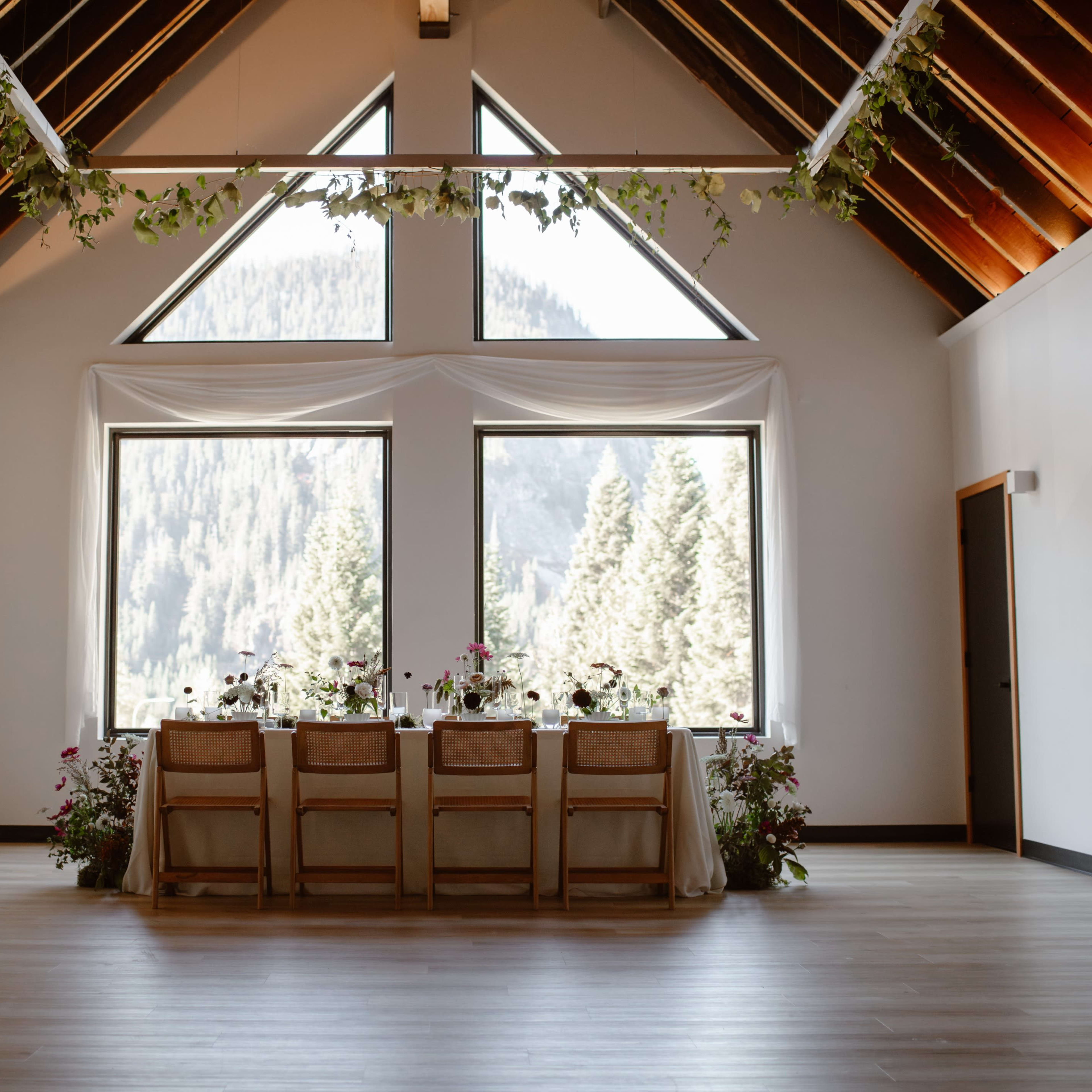 A long dining table with floral arrangements is set against large windows that showcase a forested mountain landscape.