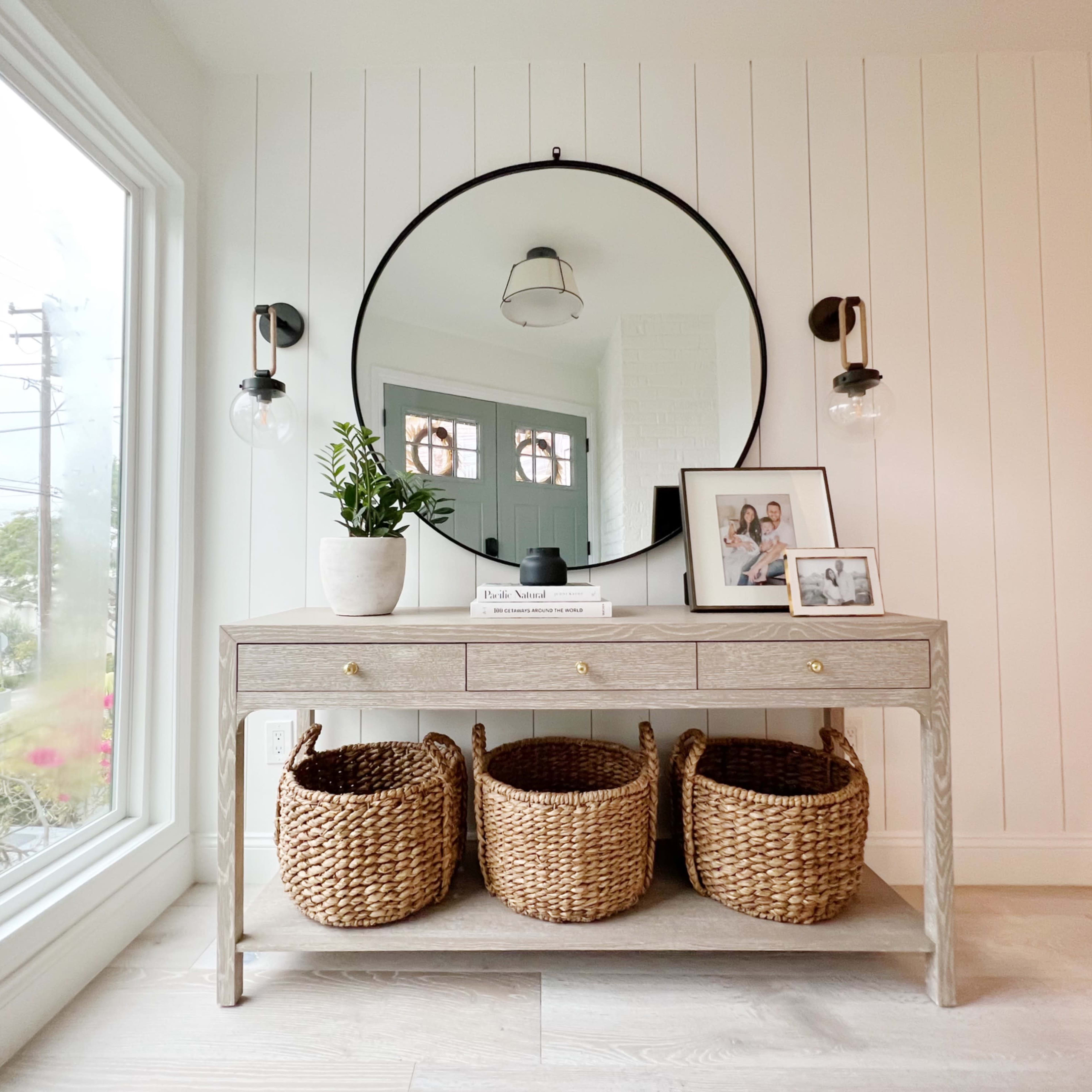 A light-colored console table with three woven baskets and a round mirror is positioned against a paneled wall near a large window.