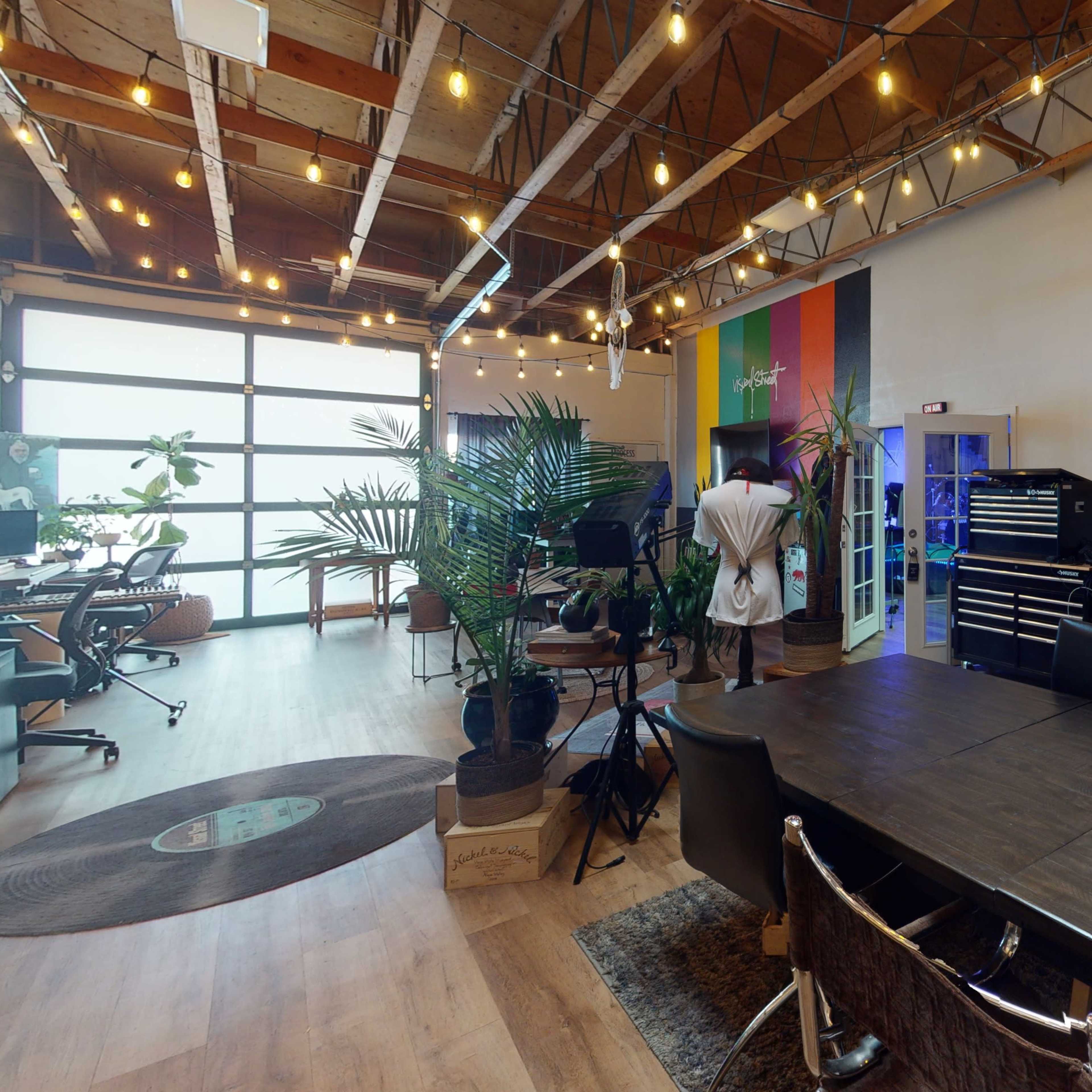 A spacious office-like environment with a large wooden table, chairs, shelves filled with equipment, and plants, illuminated by string lights.