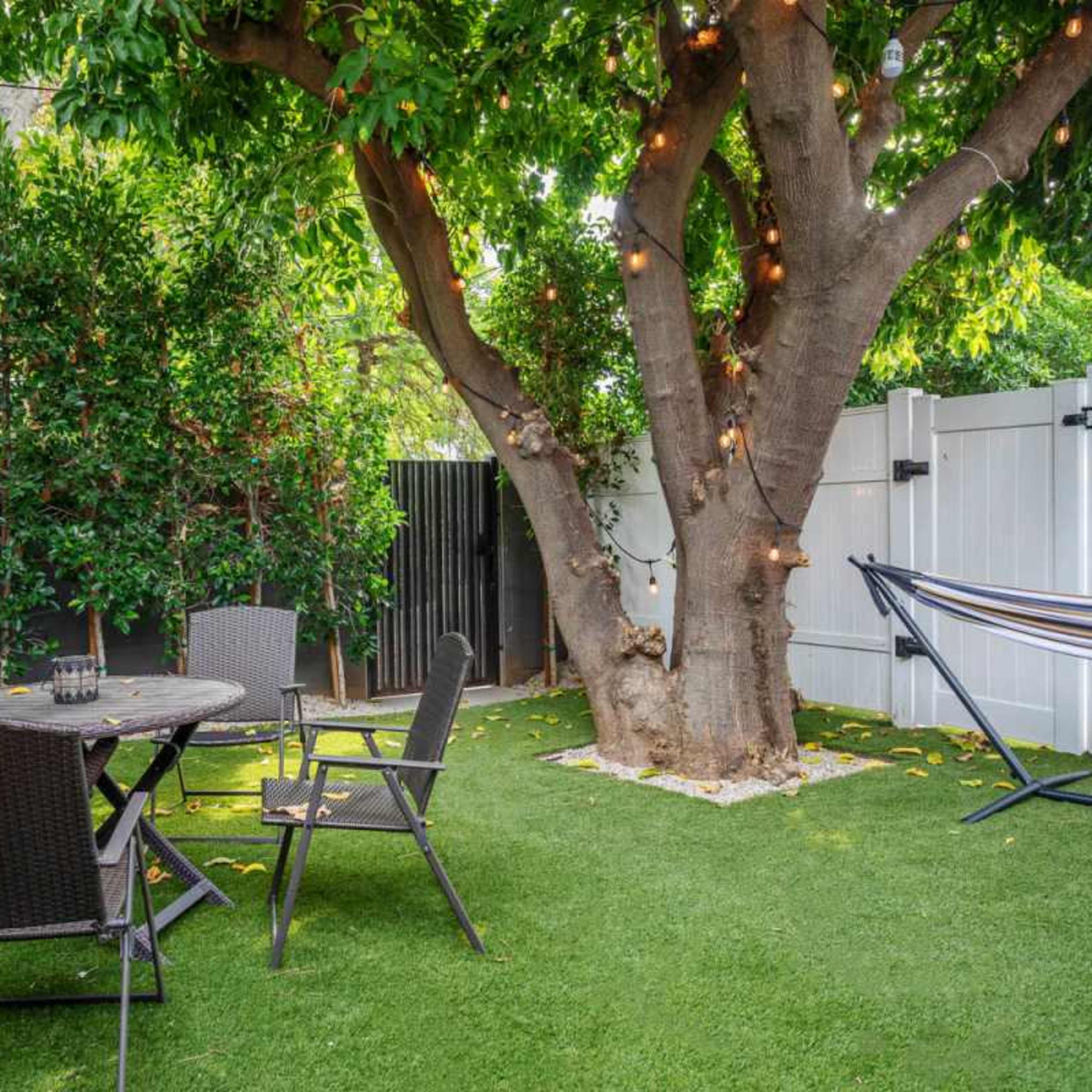 A peaceful backyard featuring a round table with four chairs, a hammock, and a large tree adorned with lights.