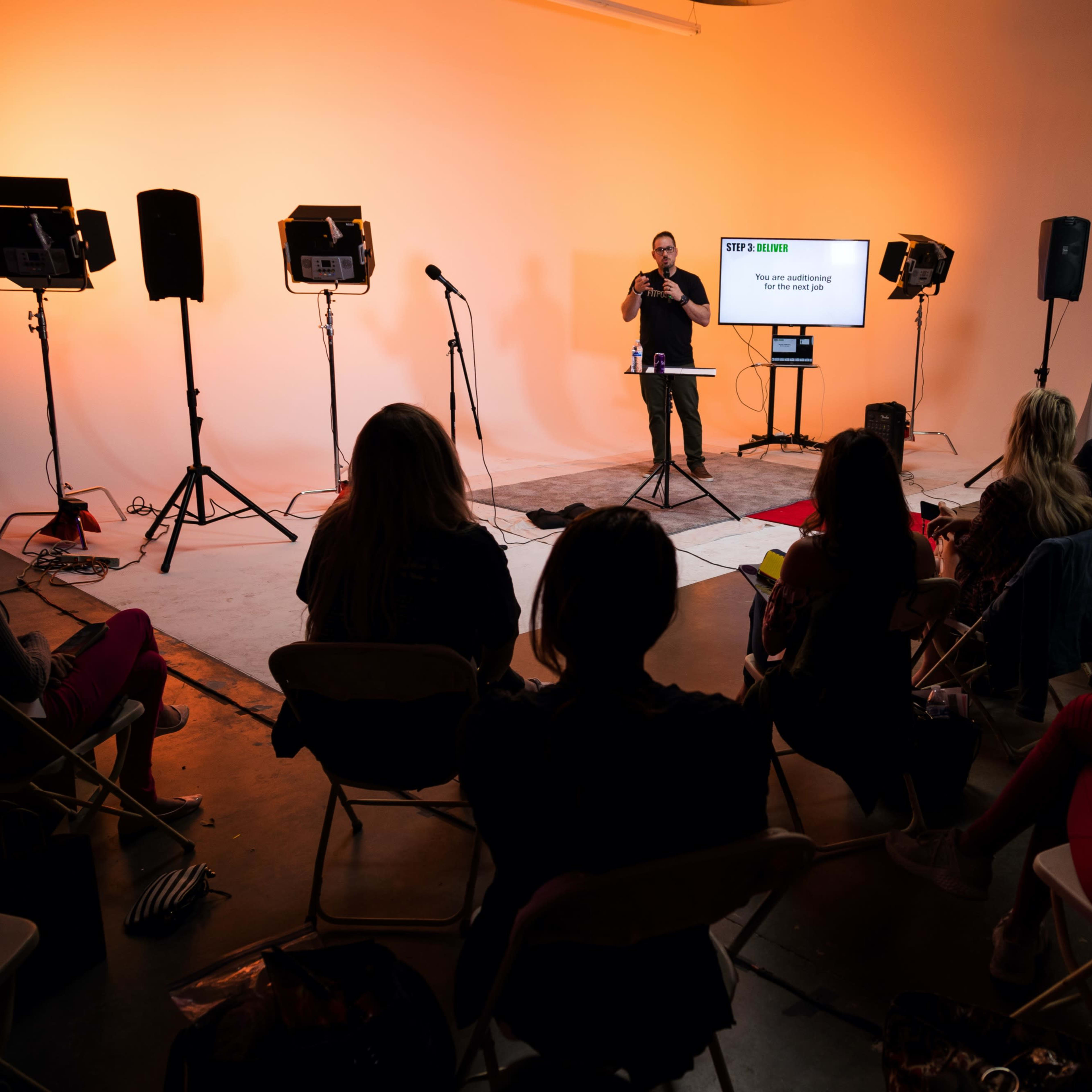 A speaker presents in front of an audience in a studio lit with warm orange lighting.