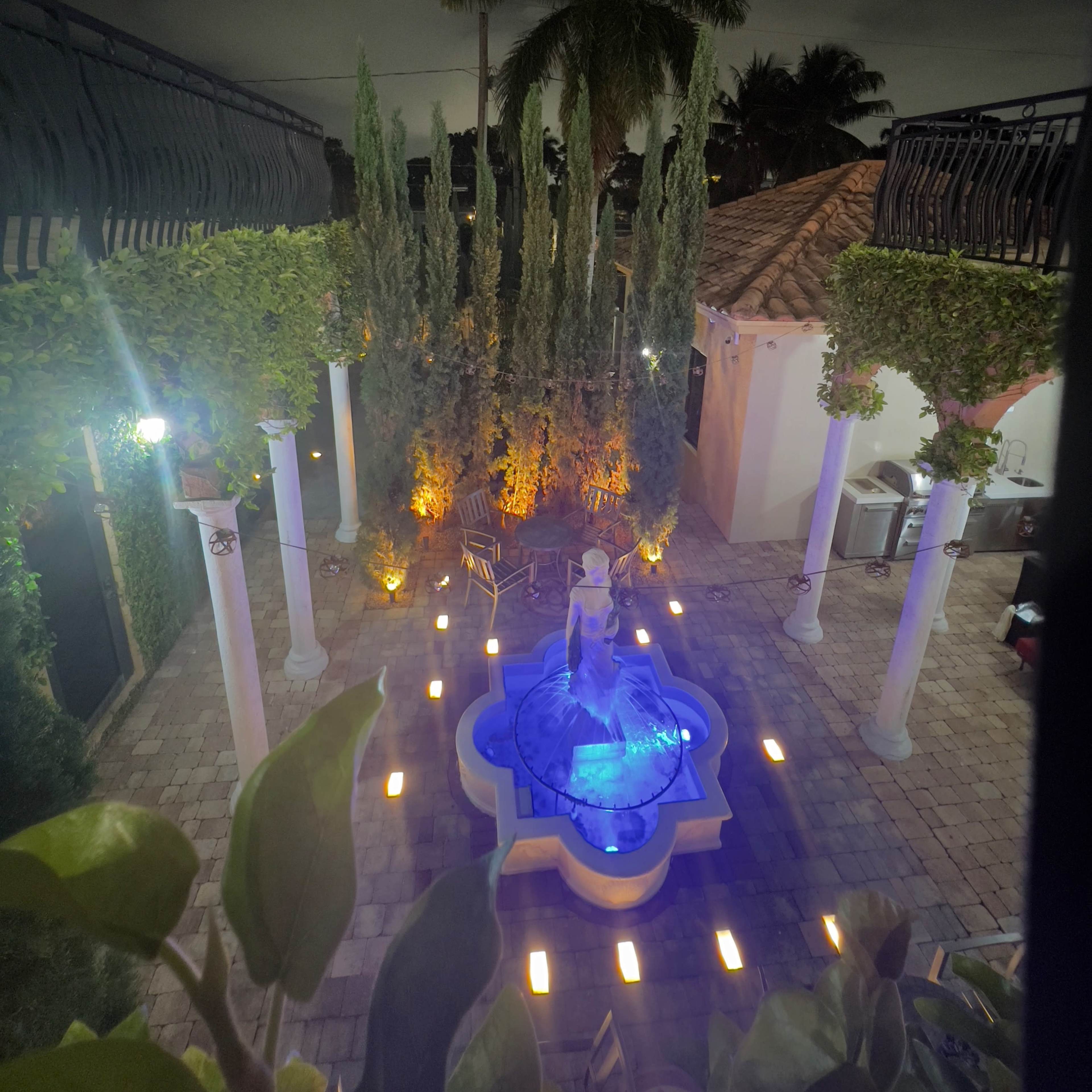 A night view of a courtyard featuring a fountain at the center, surrounded by tall greenery and illuminated by soft lights.