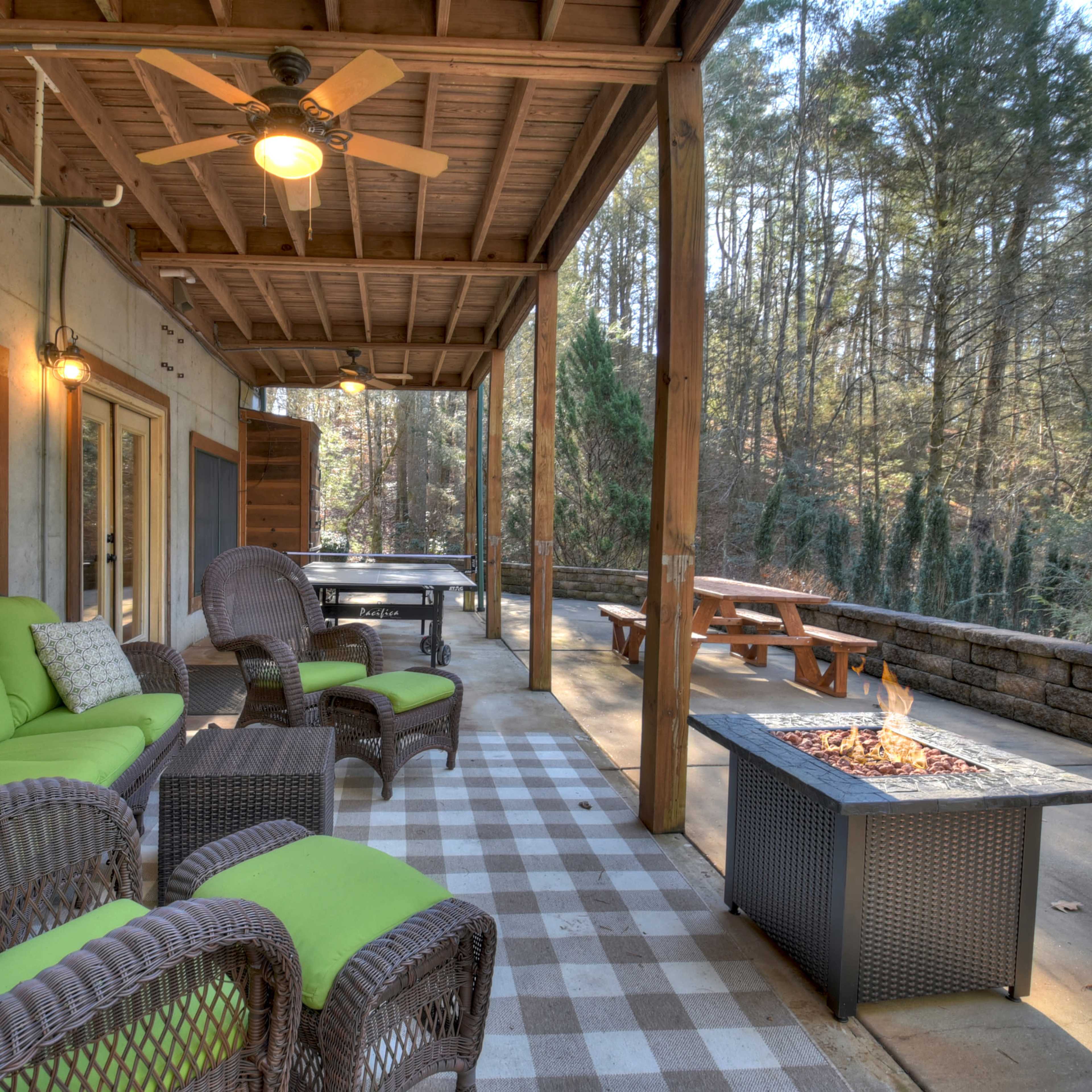 A covered patio with wicker furniture, a fire pit, and a picnic table, surrounded by trees.