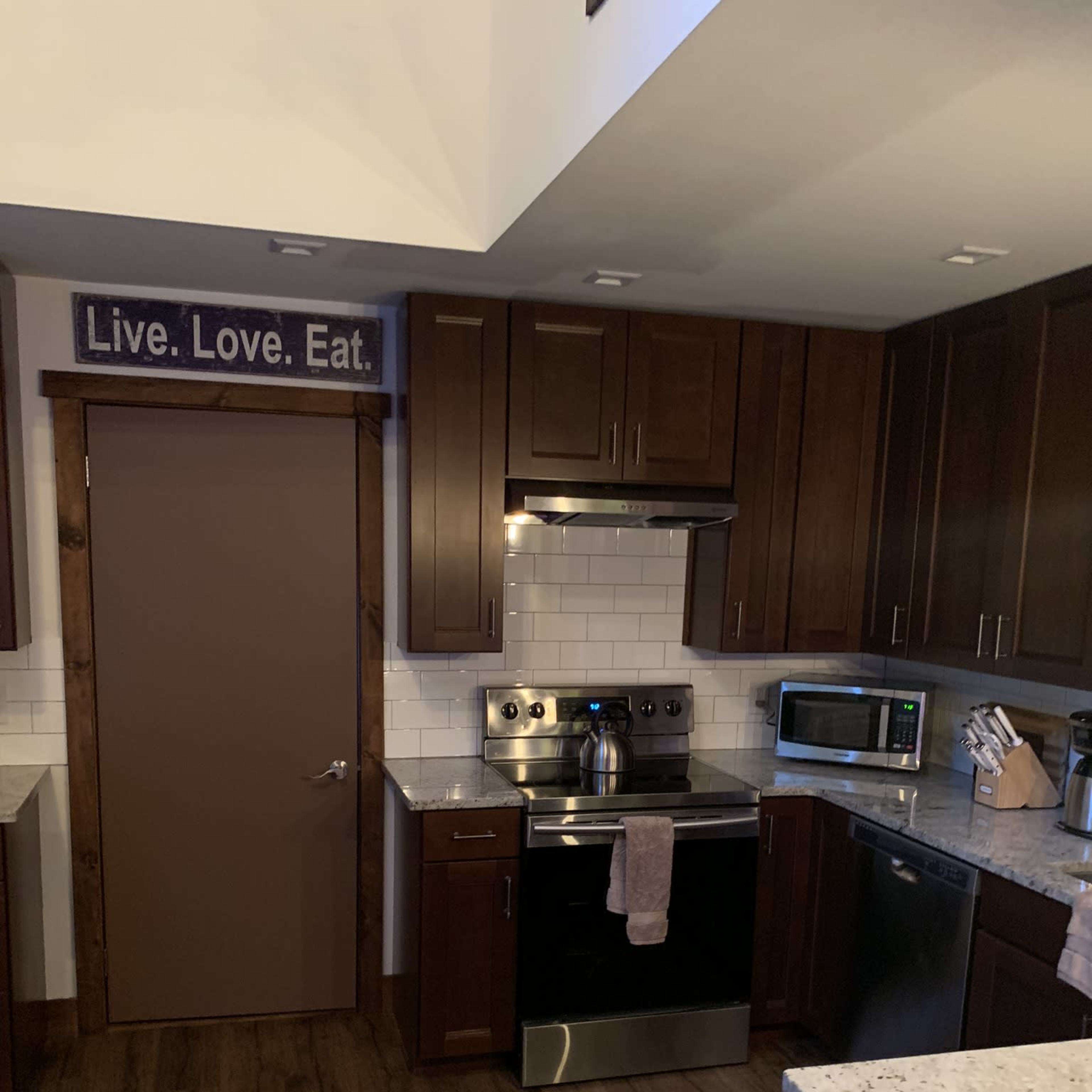 A modern kitchen features dark wooden cabinets, a stainless steel stove, a microwave, and granite countertops, with a sign reading "Live. Love. Eat." on the wall.