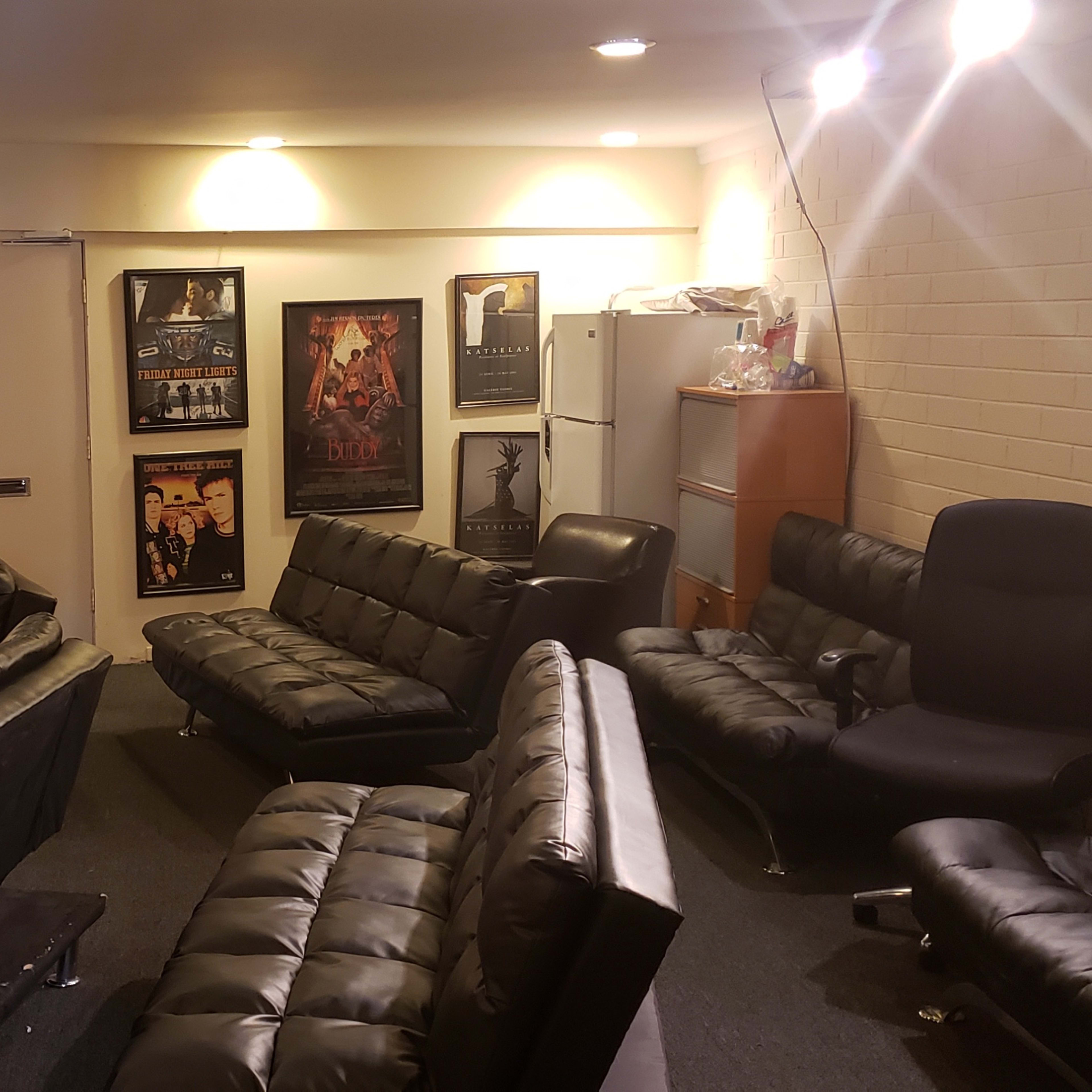 A small, dimly lit room furnished with black leather couches arranged around a central area, with posters displayed on the walls and a table on one side.