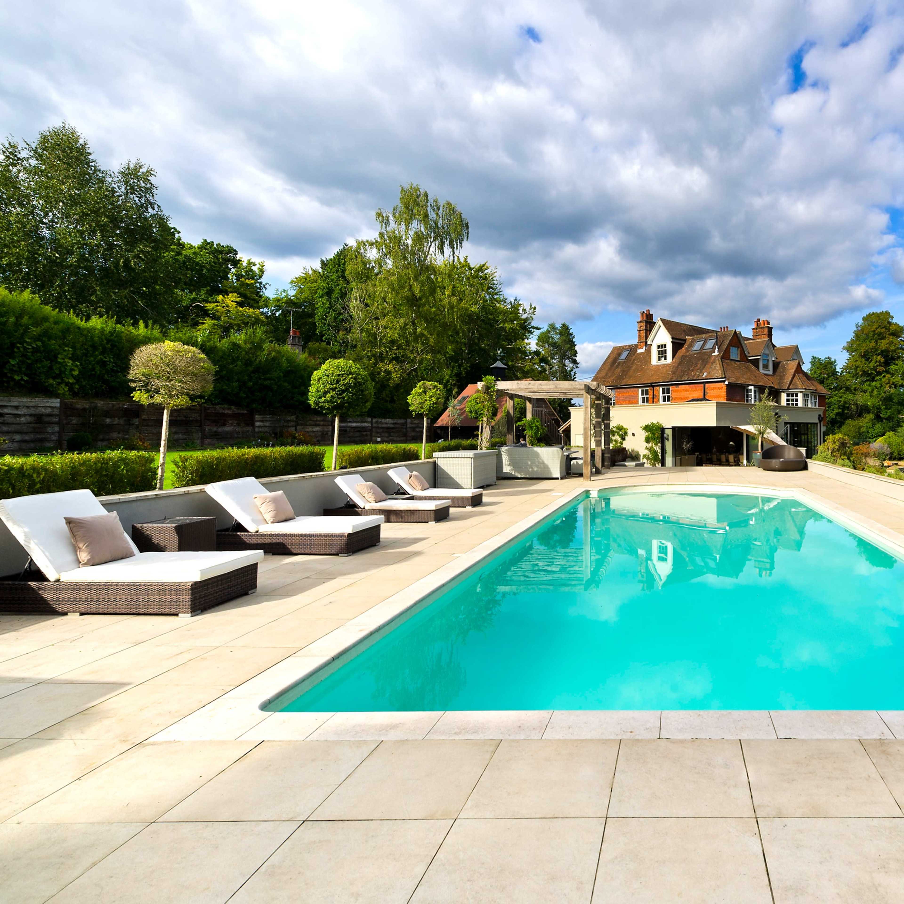 A rectangular swimming pool is surrounded by lounge chairs and overlooks a large house set against a backdrop of trees and cloudy skies.