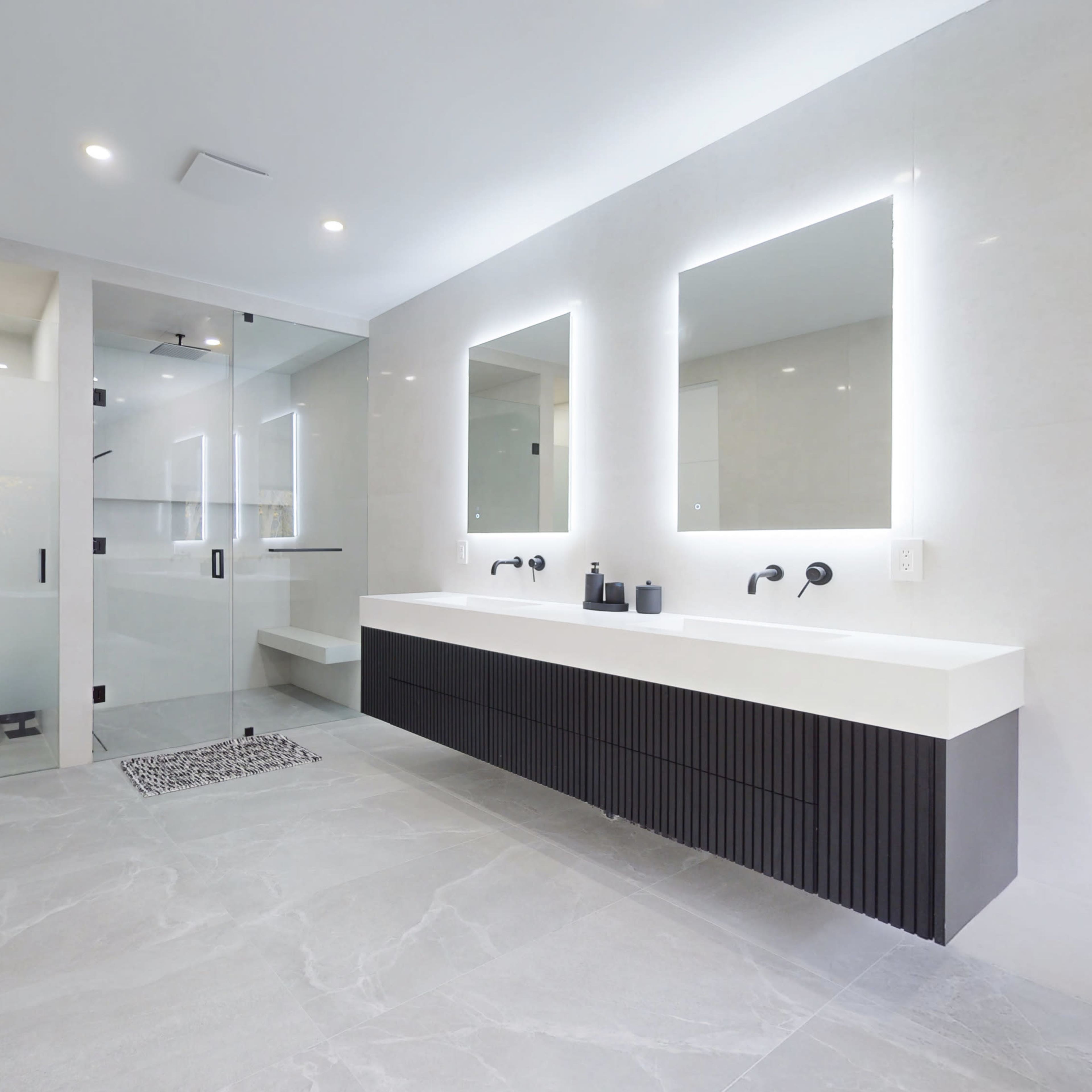A modern bathroom featuring double sinks with illuminated mirrors, a glass shower enclosure, and a minimalist design with gray tile flooring.