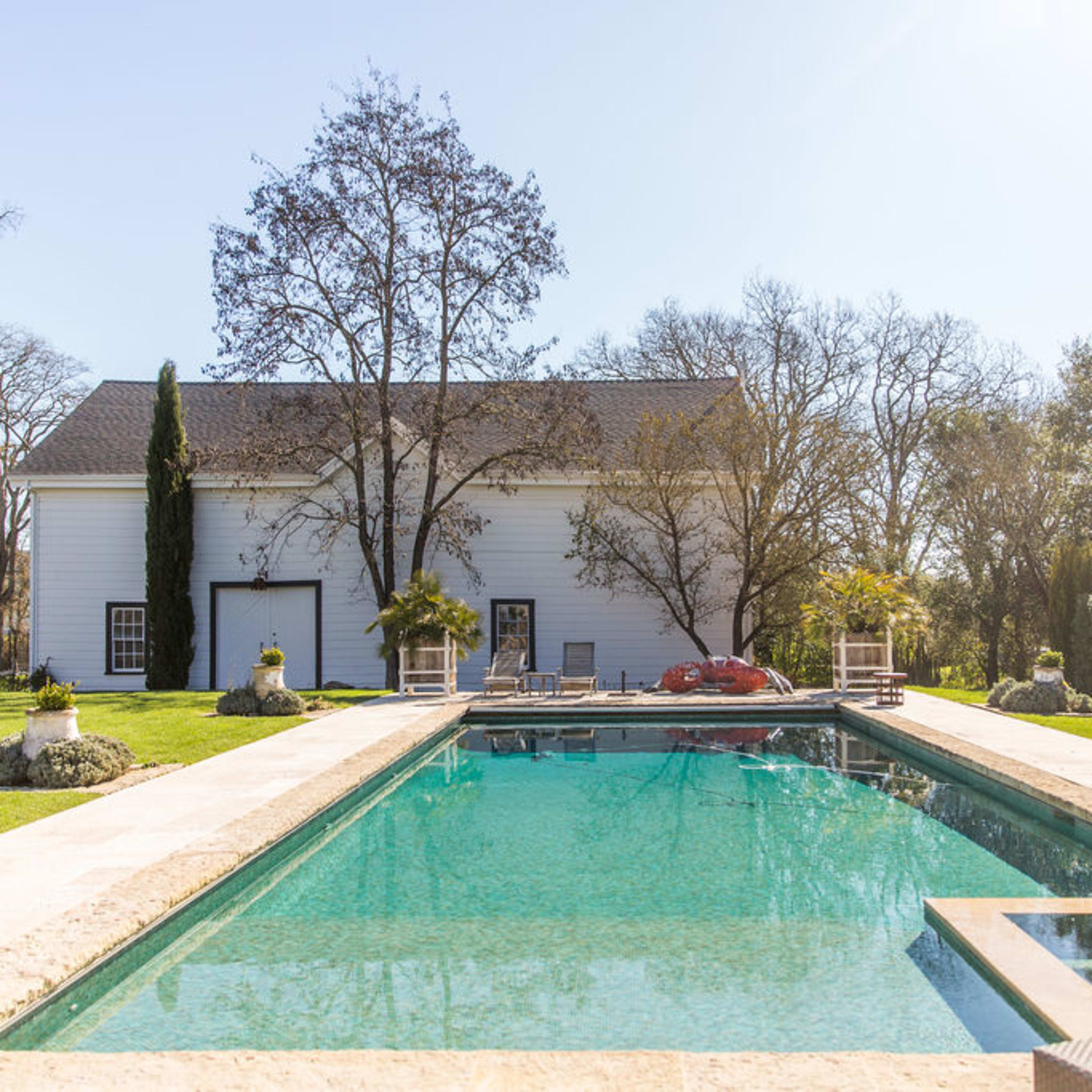 A clear rectangular swimming pool set in a garden with a white house and trees in the background.