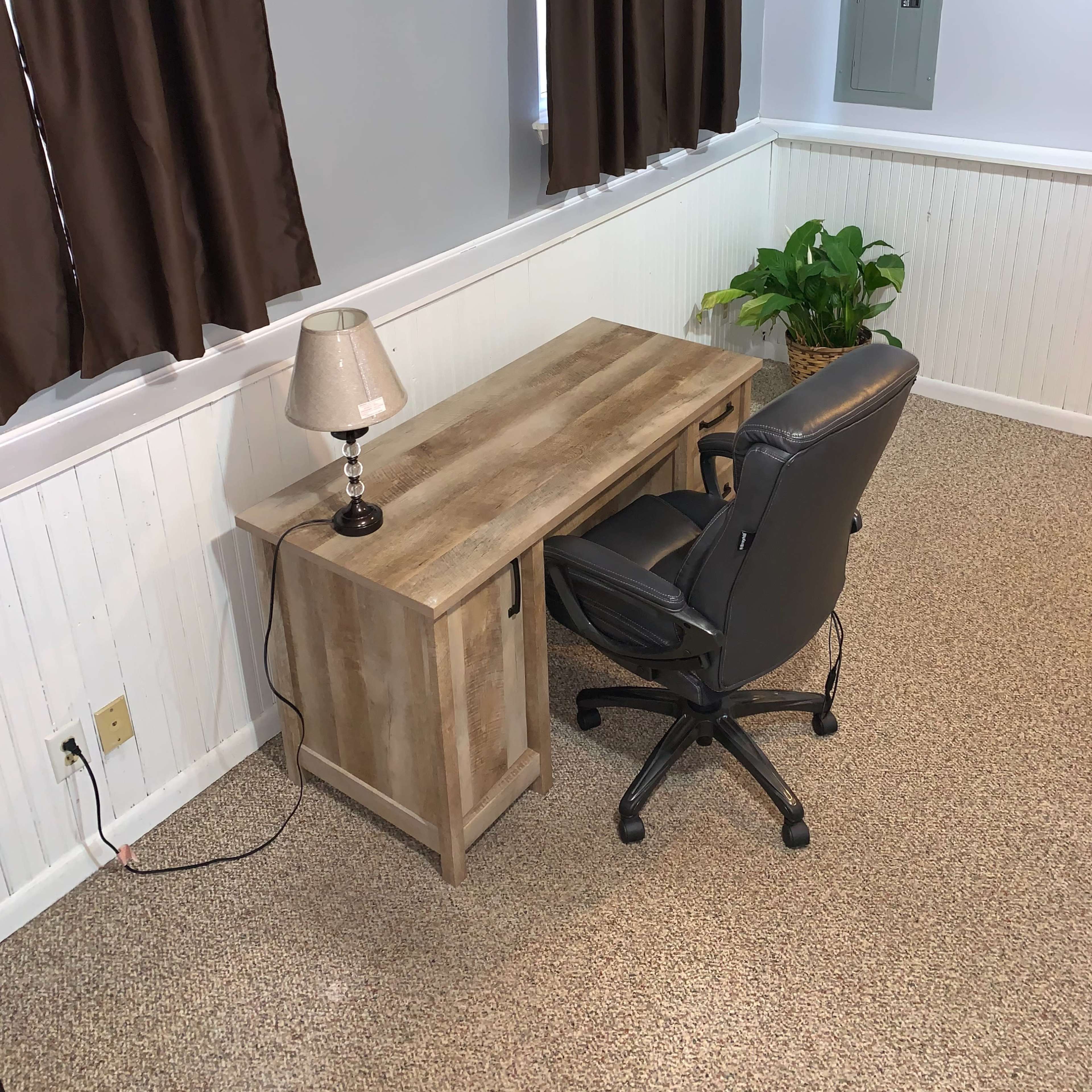 A simple home office setup with a wooden desk, an office chair, a lamp, and a potted plant, all situated in a room with brown curtains and beige carpet.