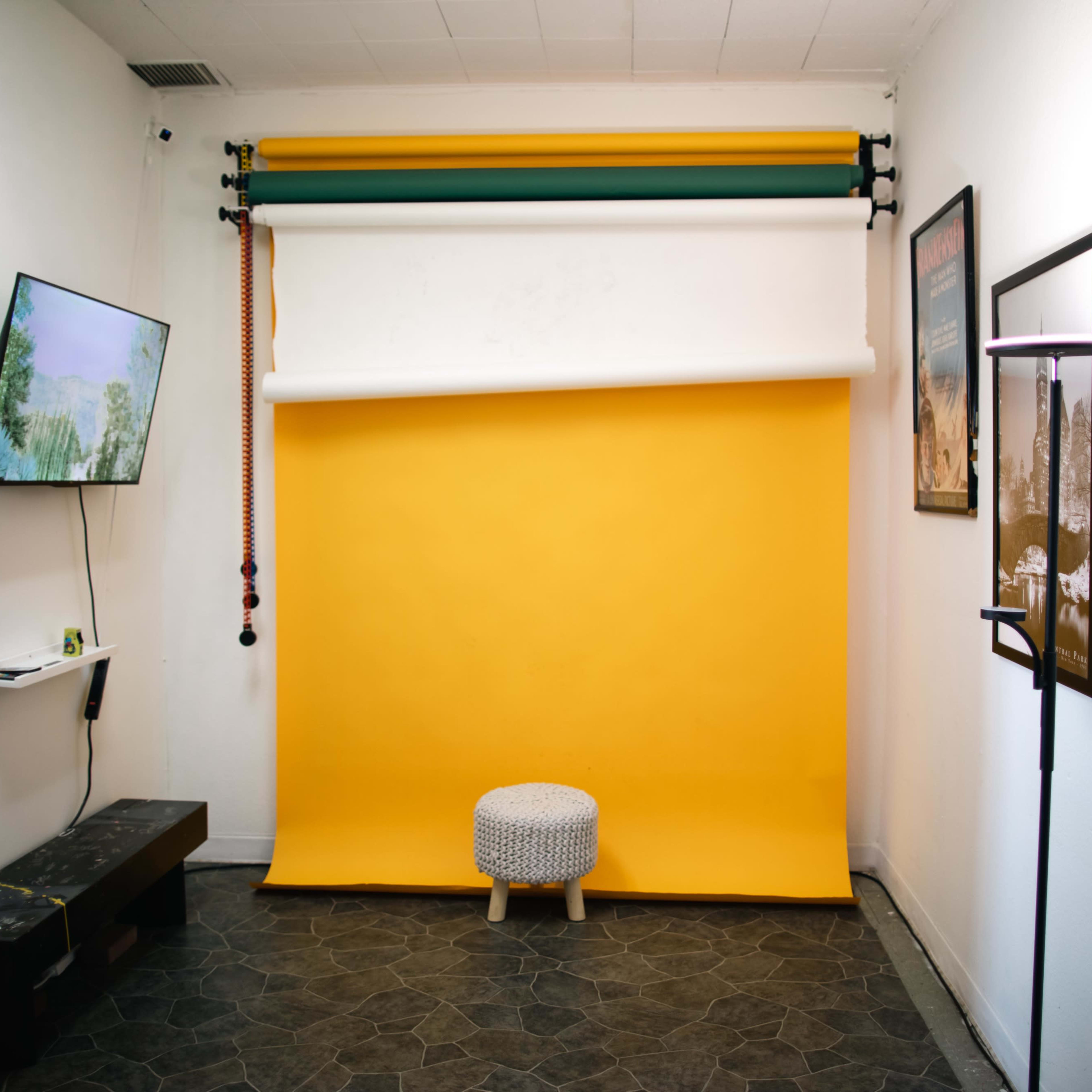 A small studio space featuring a yellow backdrop, a stool, and a television mounted on the wall.