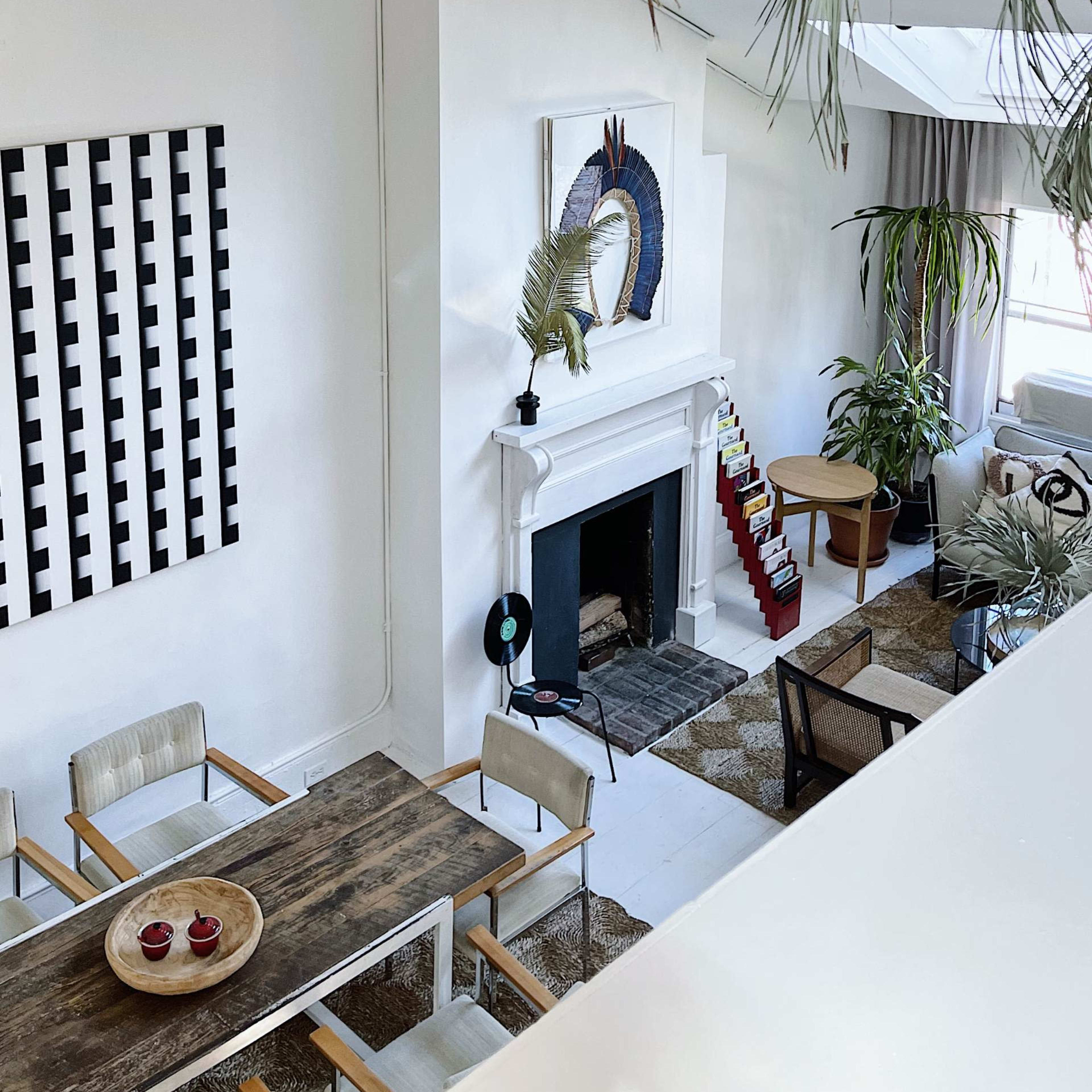 A modern living room with a white fireplace, a dining table, and large plants, accented by a geometric wall art piece.