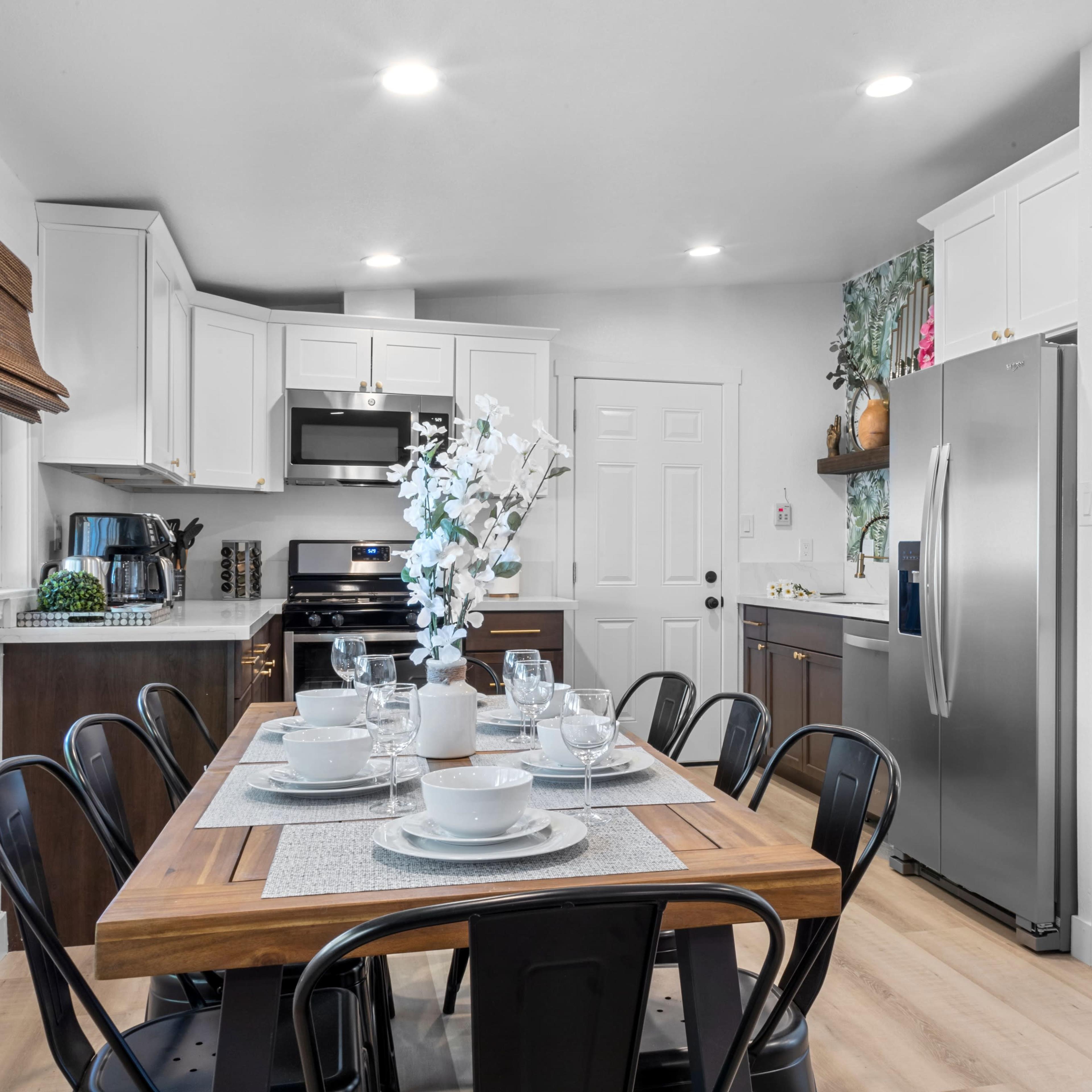 A modern kitchen features a wooden dining table set for eight with white dinnerware, surrounded by black chairs, and equipped with stainless steel appliances and white cabinetry.